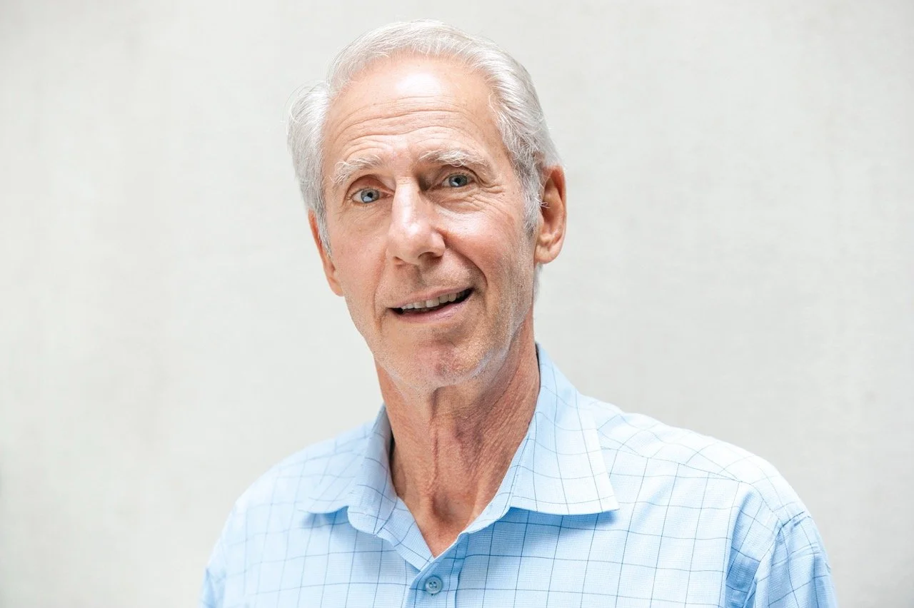 A smiling elderly man with gray hair, wearing a light blue checkered shirt, standing against a plain off-white background.