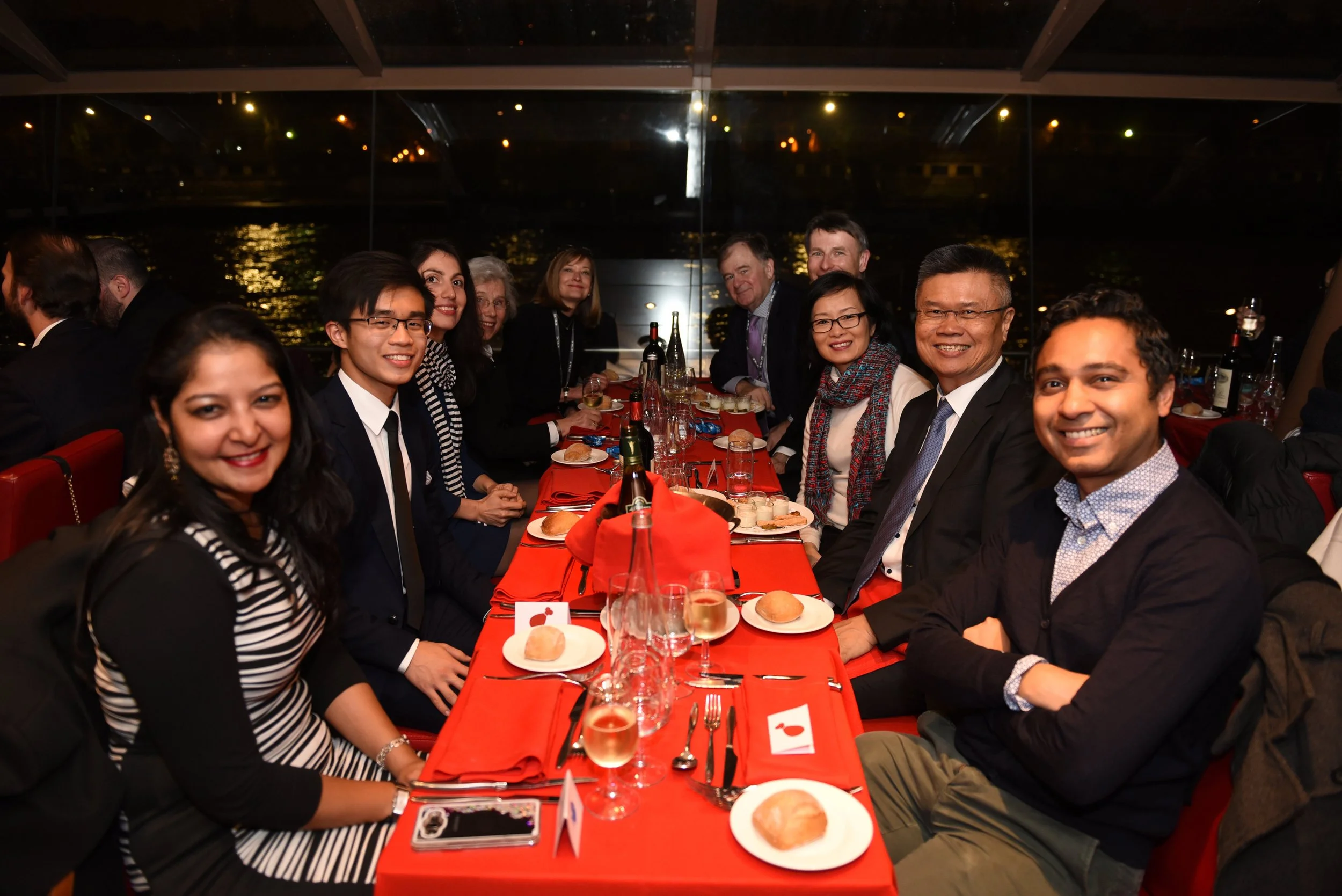 People gathered around a dining table at a dinner event, with a city skyline visible through large windows in the background.