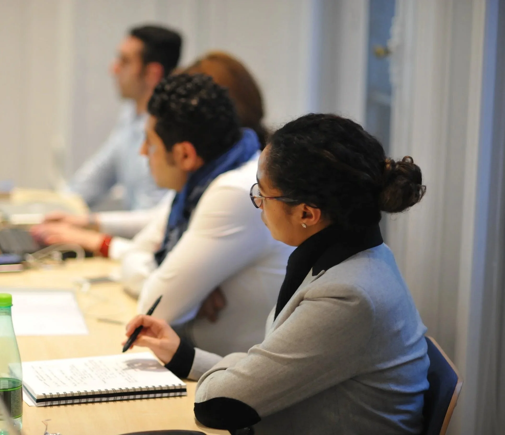 Four people sitting at a conference table in a meeting room, with notebooks and pens in front of them, attentively listening.