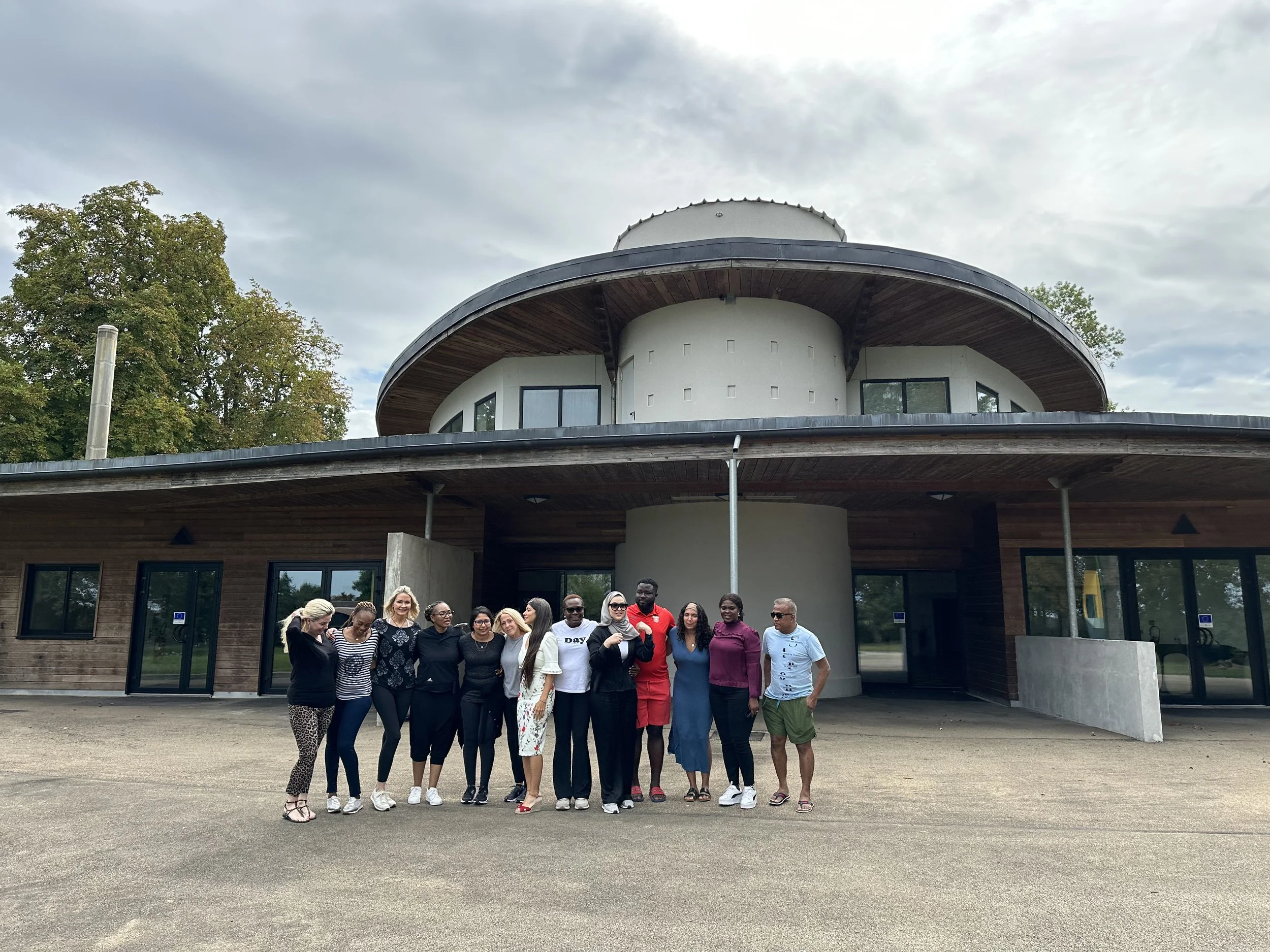 Group of people standing outside in front of a modern, round building with large windows and a wooden exterior.