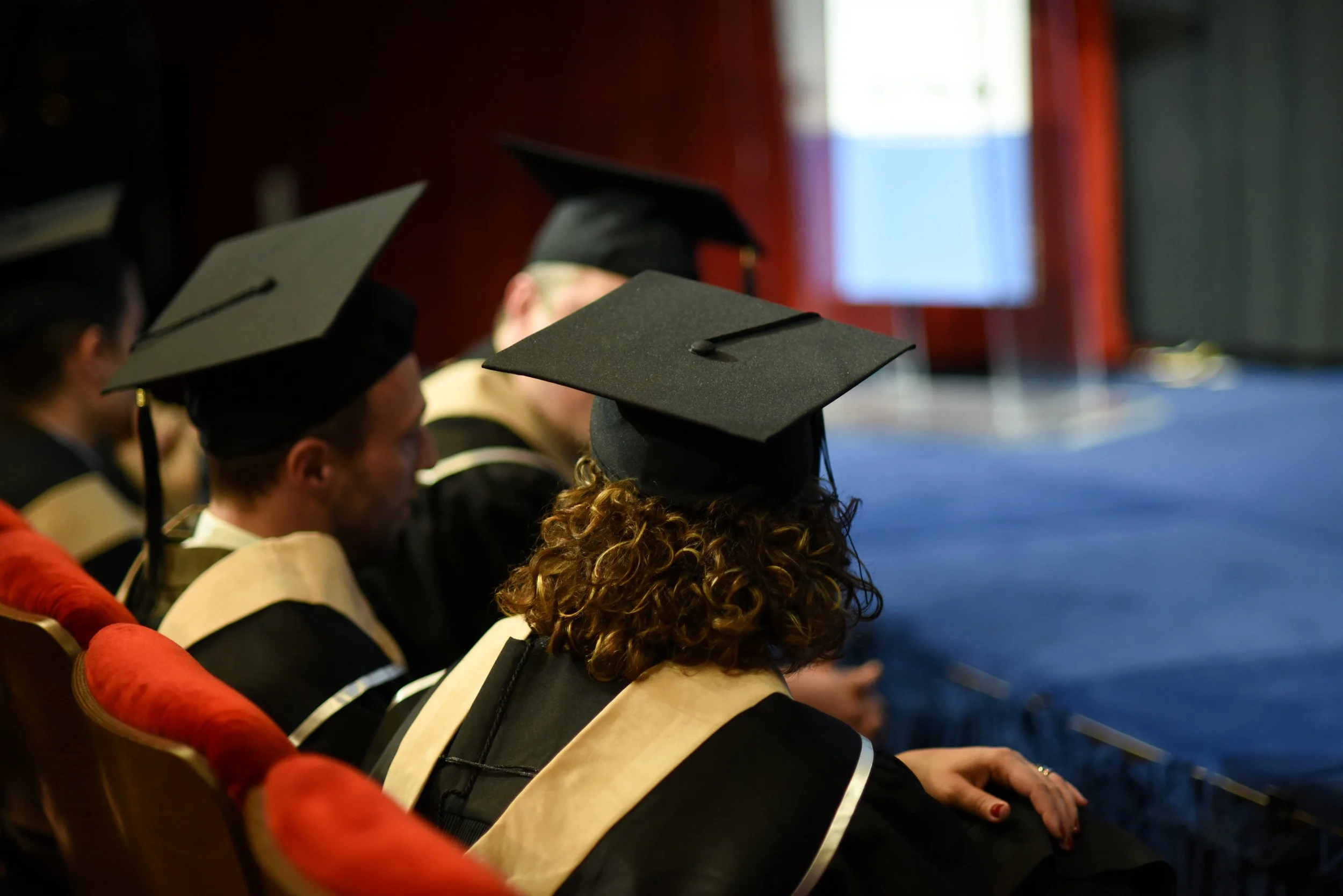 People in graduation caps and gowns attending a graduation ceremony, sitting in red chairs facing a stage with a blue carpet.