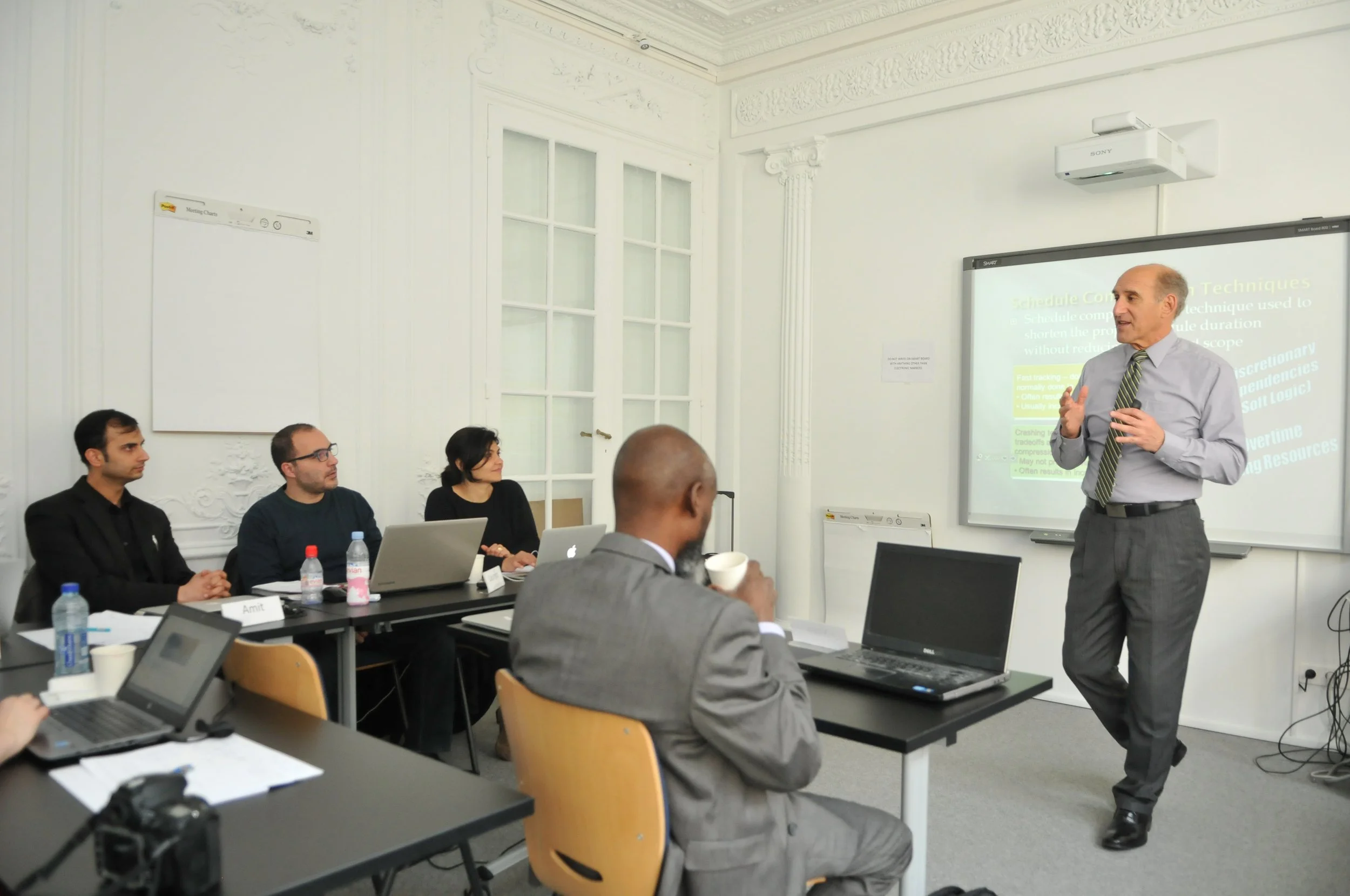 A professional man giving a presentation to a group of people in a conference room with white walls and large windows, using a digital projector and screen.