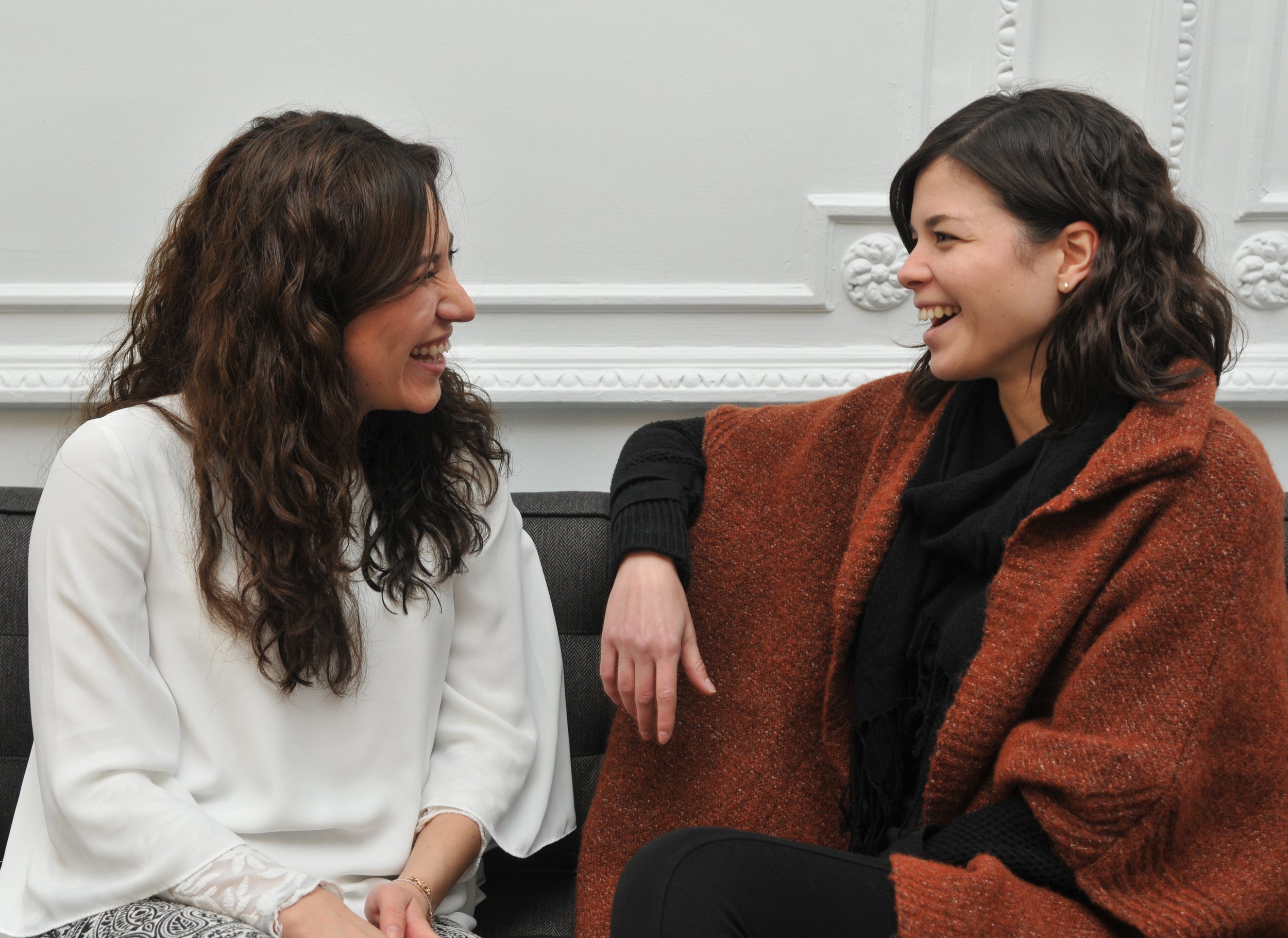 Two women sitting and laughing together, one wearing a white top and the other wearing a brown coat with black top underneath.