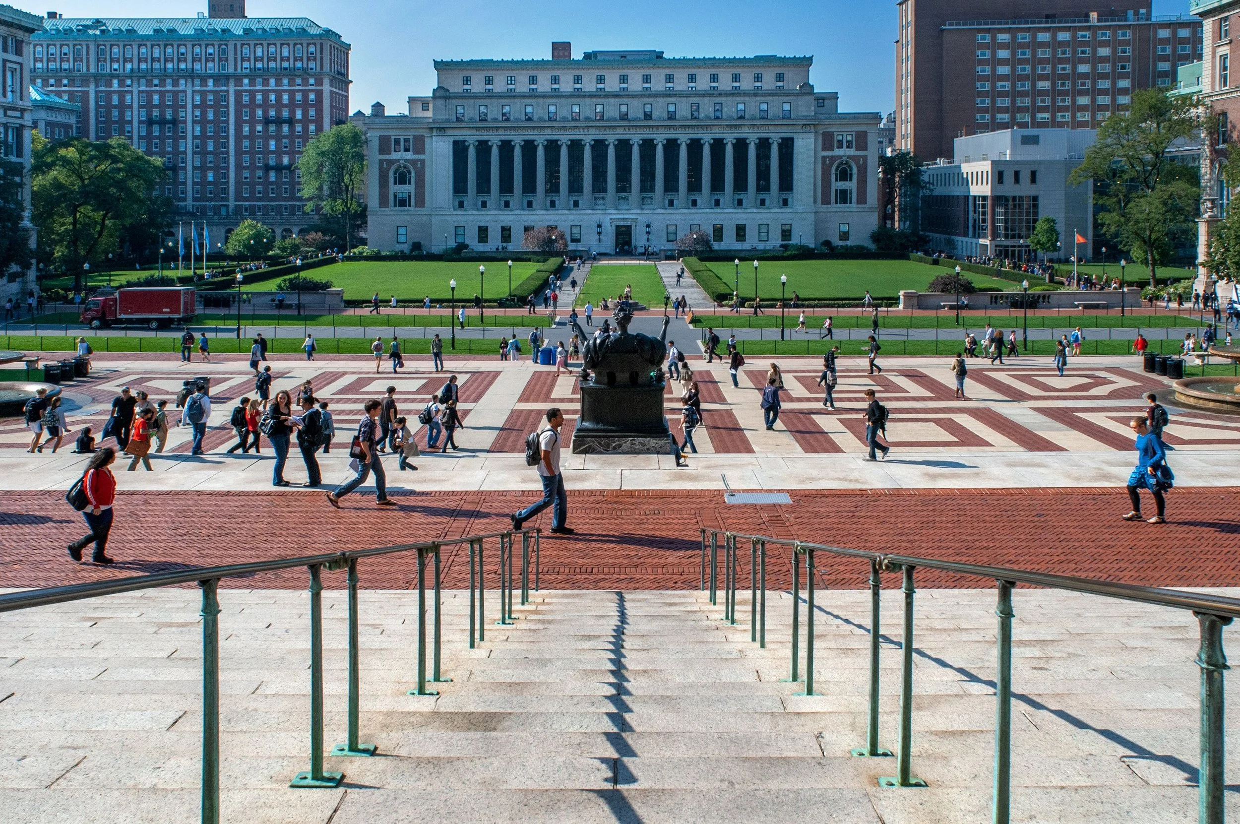 People walking across a historic university campus with a large building, green lawns, and a fountain in the background.