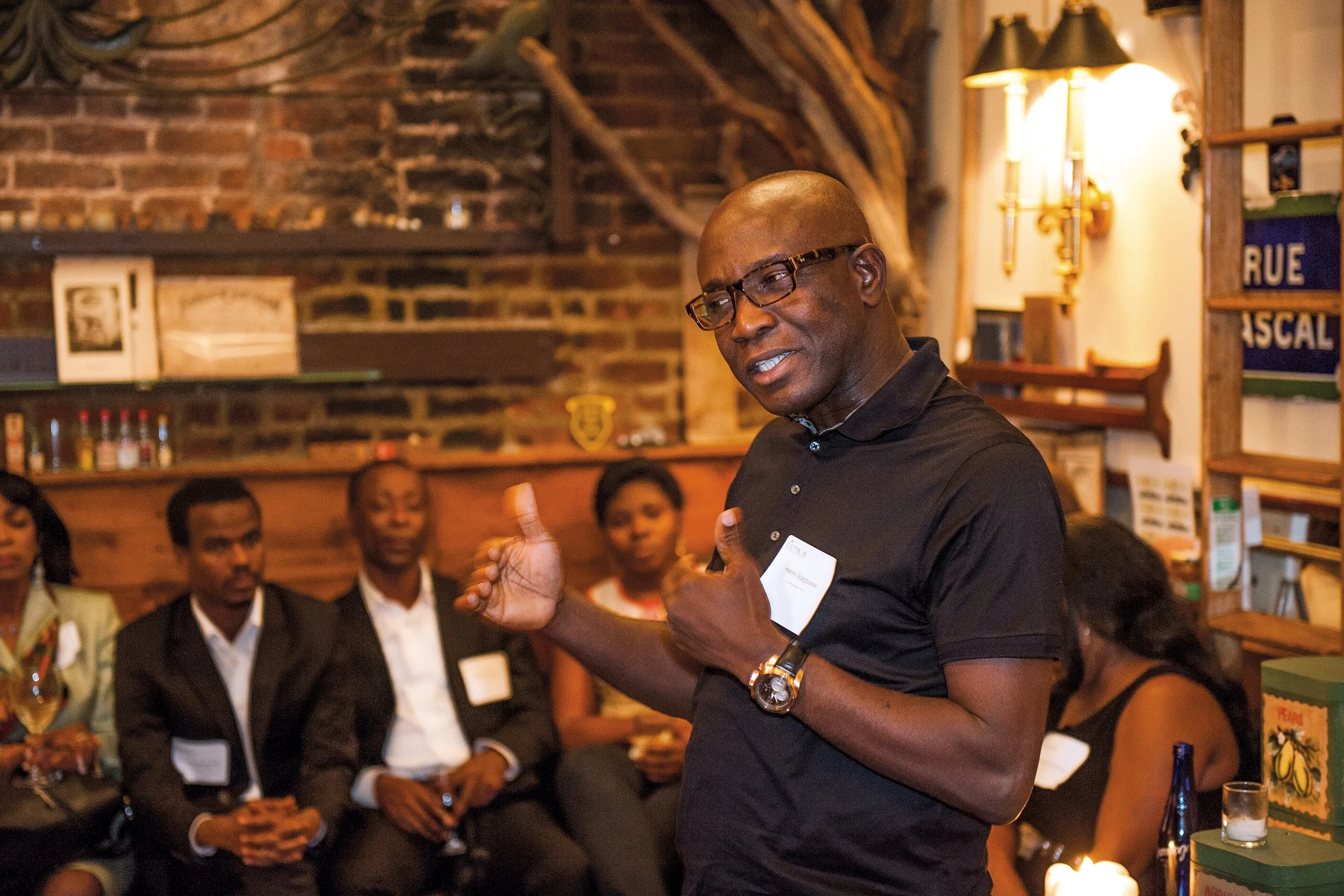 A man wearing glasses and a black shirt speaking to a group of people in a cozy, wood-paneled room.