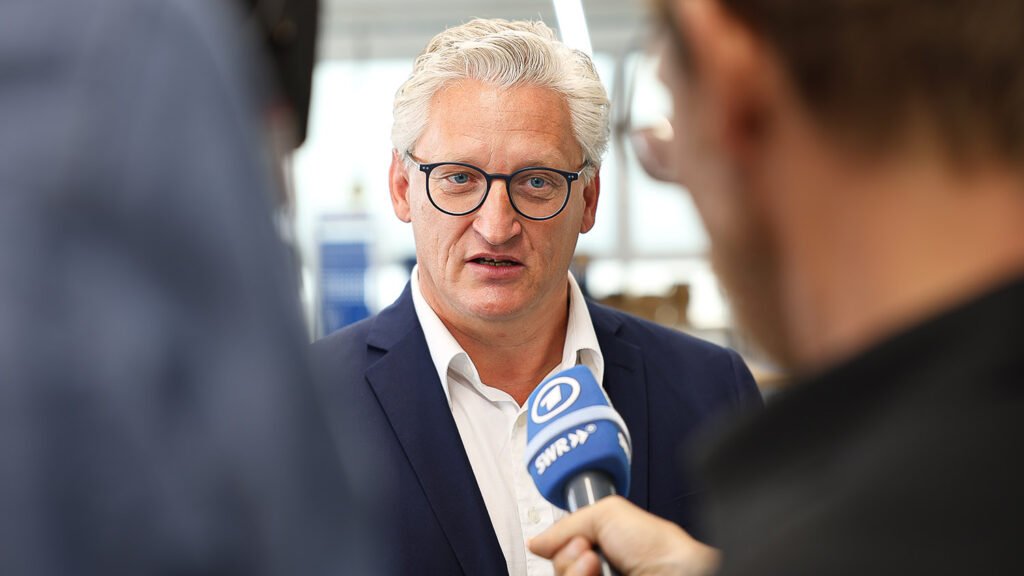 A man with white hair and glasses in a dark suit is being interviewed by a person holding a microphone with a news logo. There are two other people visible in the foreground.