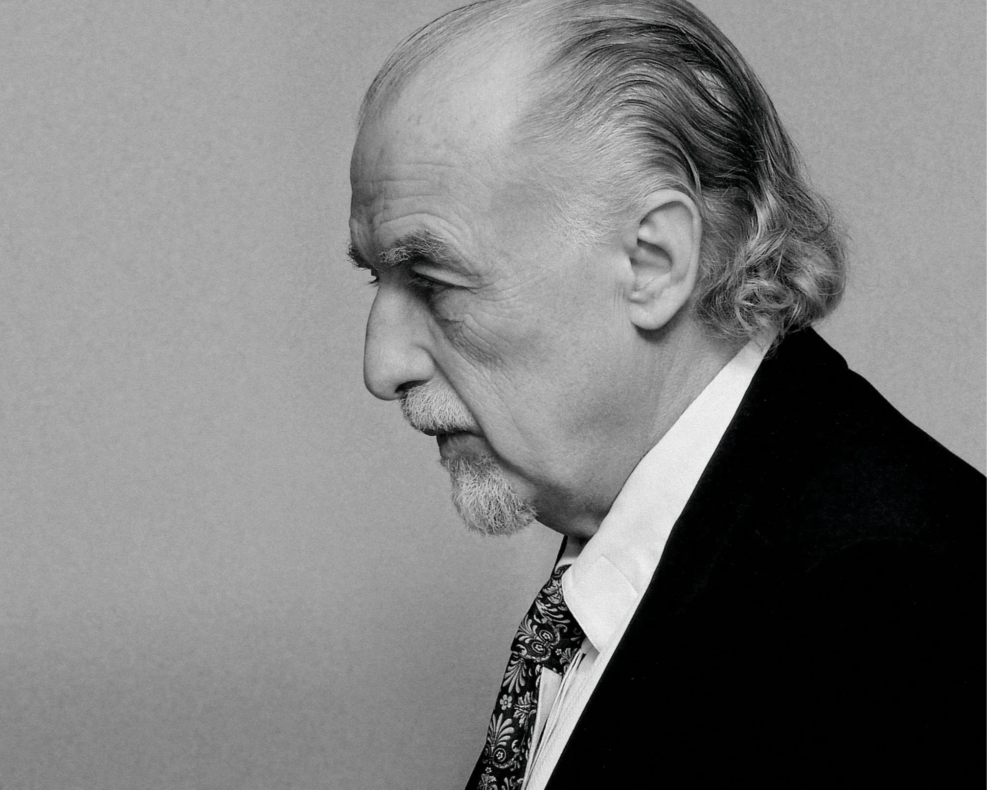 A black and white side profile portrait of an older man with a beard and slicked-back hair, wearing a suit and a patterned tie.
