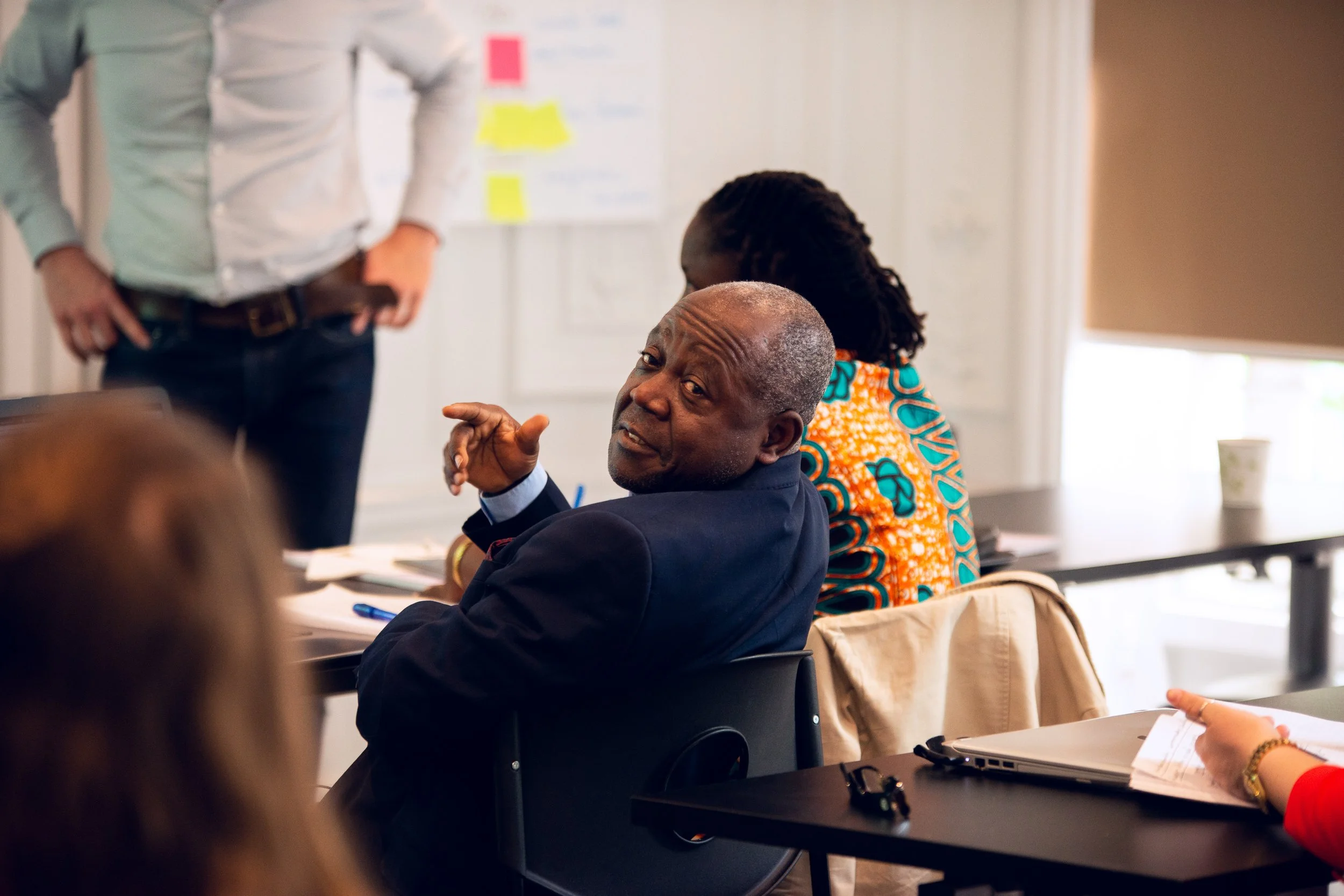 An older man in a dark suit sitting at a table in a meeting room, looking over his shoulder, engaging in conversation with others during a meeting.