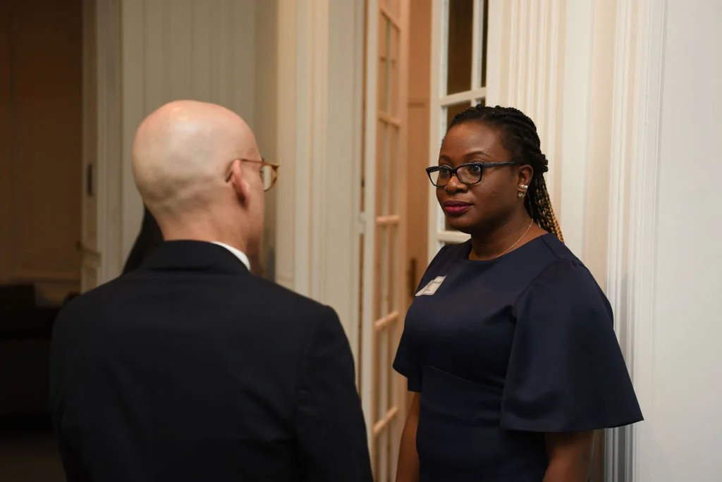 A woman with glasses and braided hair talking to a bald man with glasses in a formal setting.