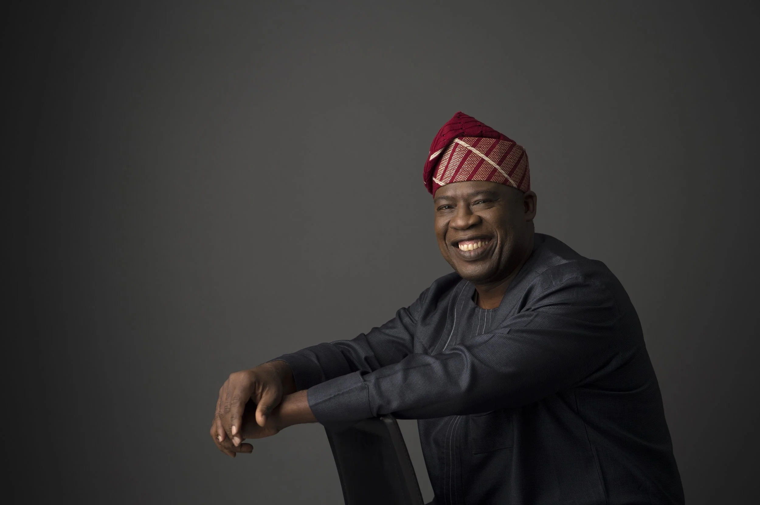 A smiling Black man wearing a traditional red and gold headwrap and a dark gray outfit, leaning on the back of a chair against a plain gray background.