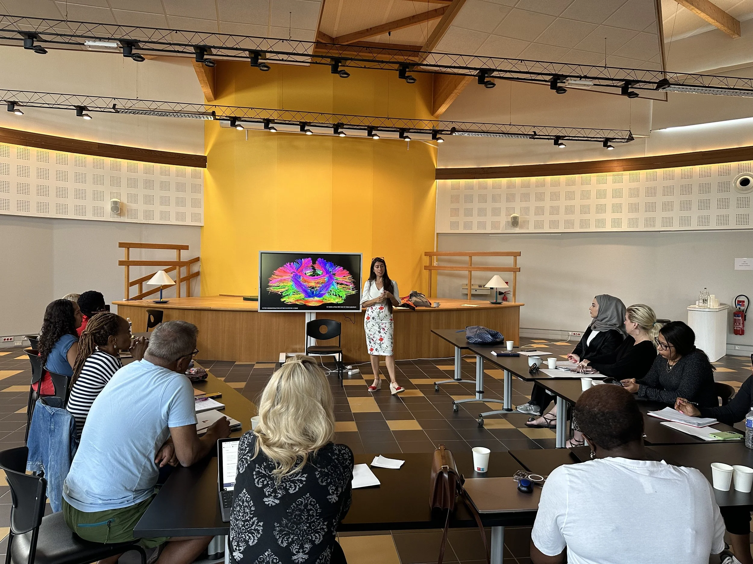 A woman giving a presentation to a group of diverse people in a conference room with yellow and gray walls, a large screen, and tables arranged in a U-shape.