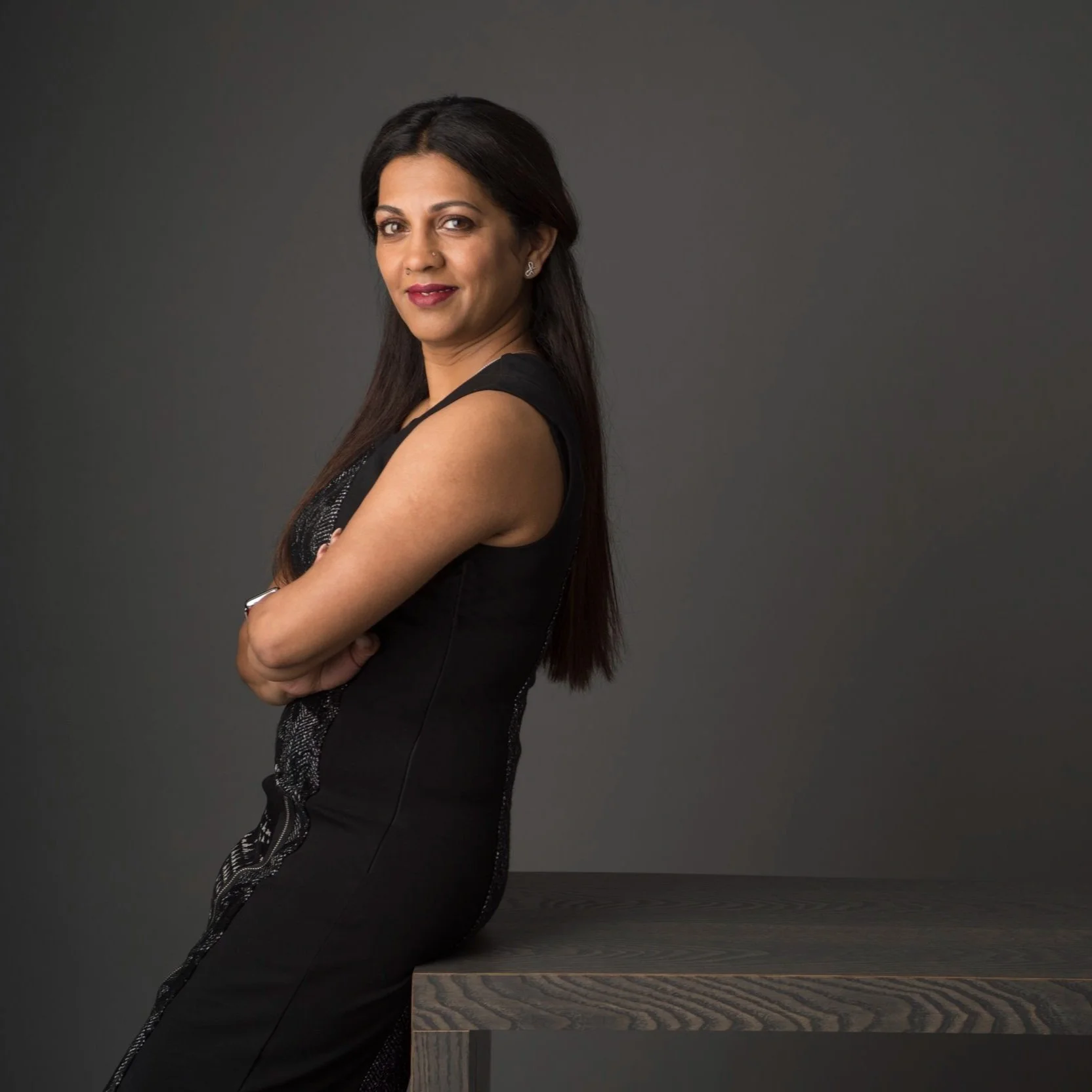 A woman with long dark hair, wearing a black sleeveless dress, sitting on a wooden table with her arms crossed against a neutral gray background.