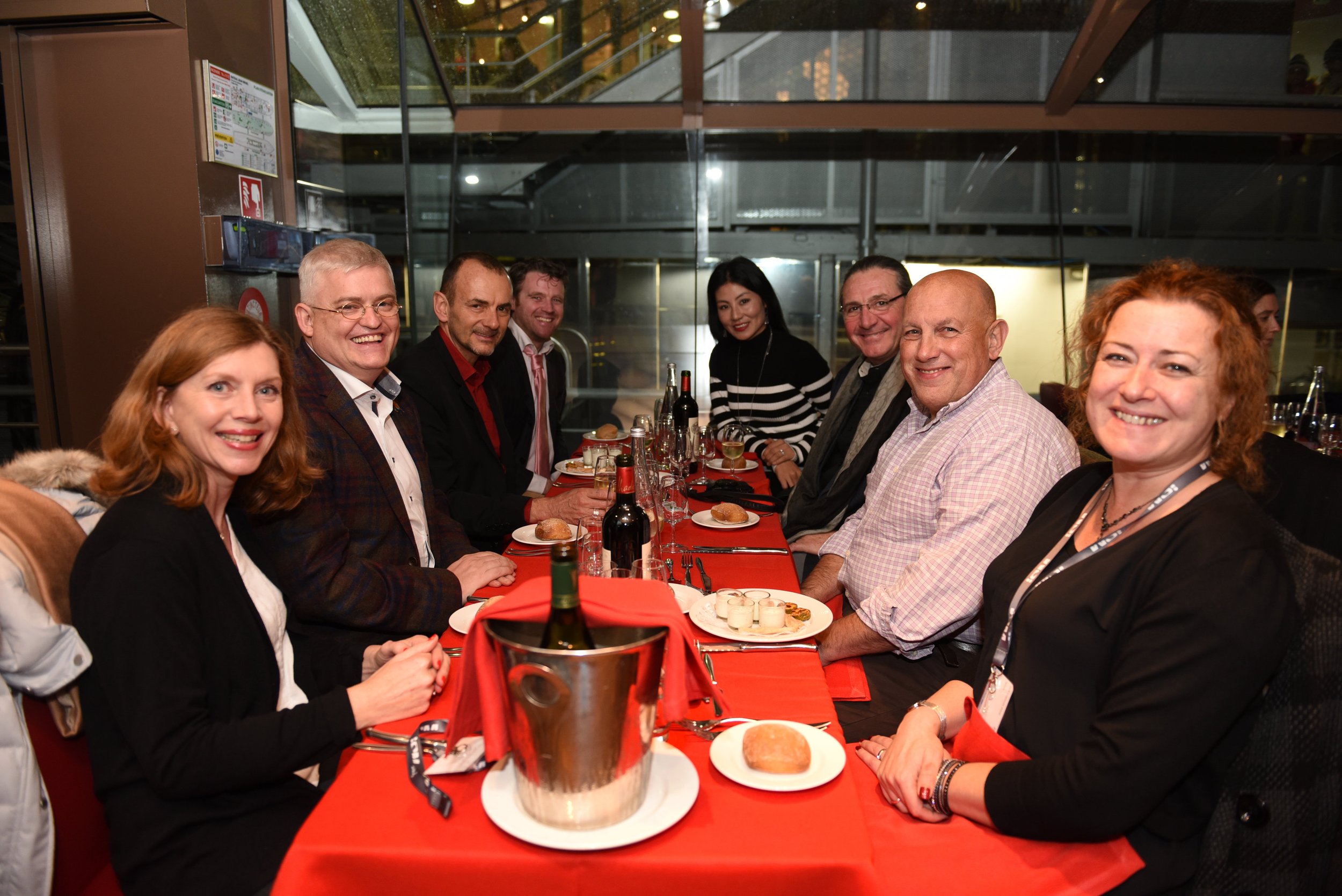 A group of nine diverse adults sitting around a dinner table, smiling at the camera, in a modern restaurant with glass windows in the background.