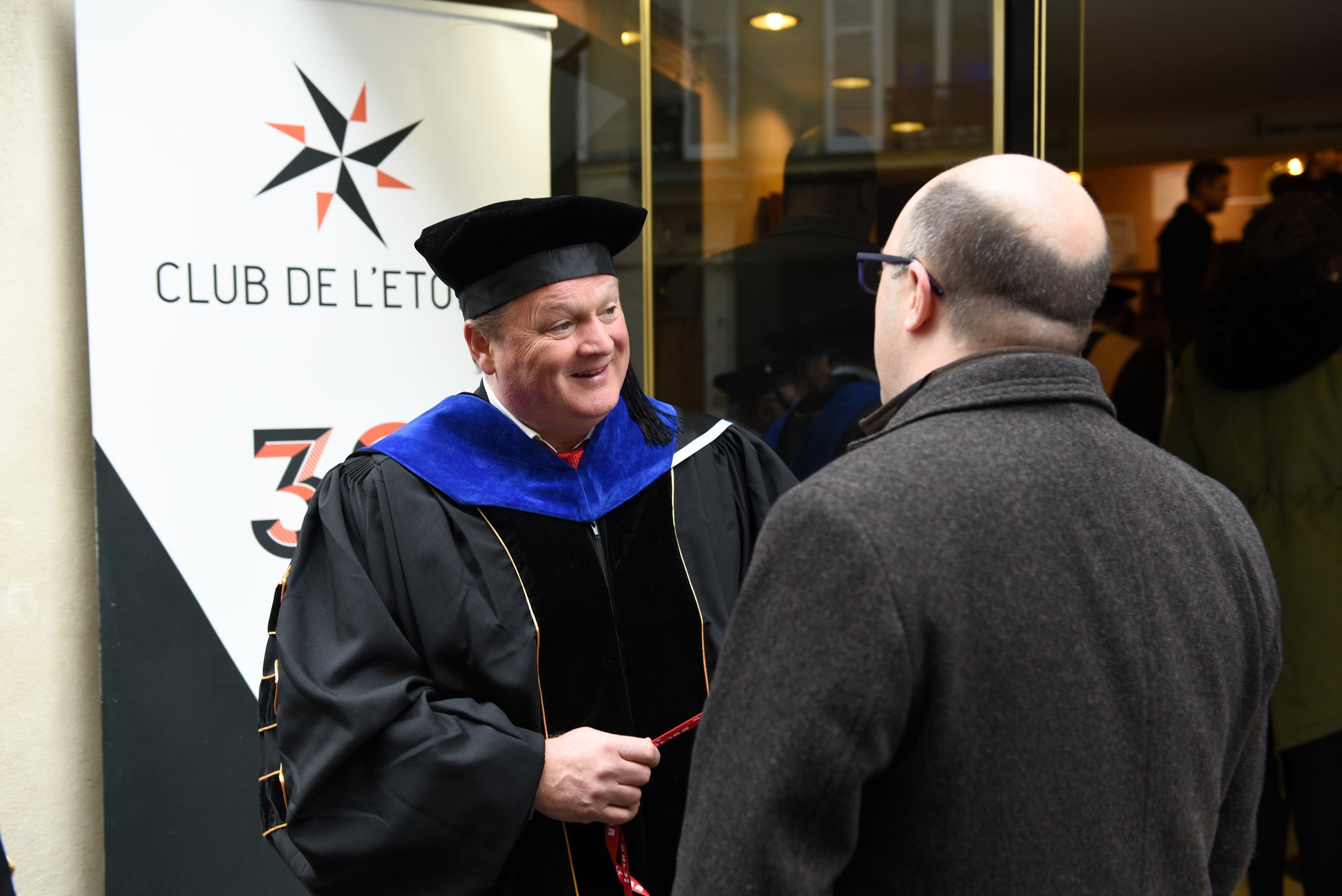 Two men engaged in conversation at a graduation event, one wearing traditional academic regalia including a black gown and cap with a blue velvet hood, standing in front of a banner that reads 'CLUB DE L'ETOILE' with a star logo.
