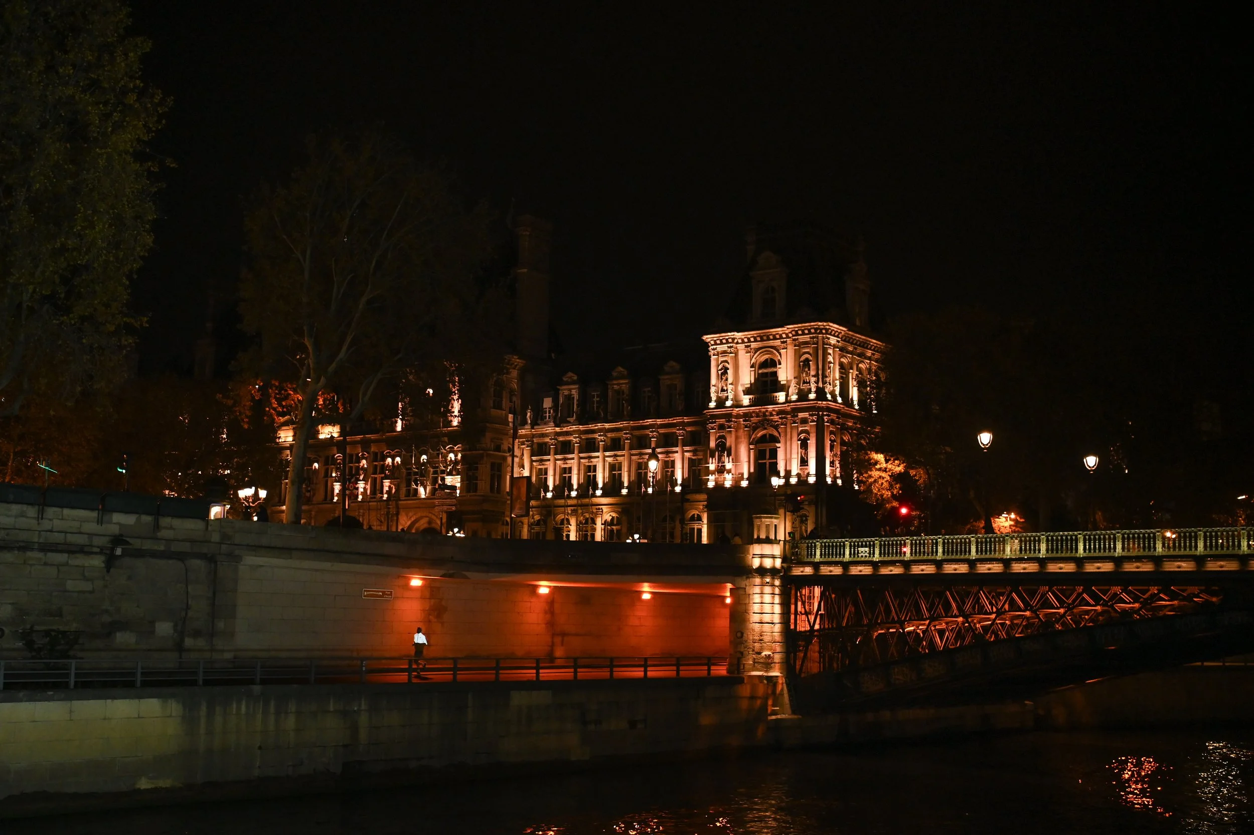 A historic building illuminated at night, situated next to a river with a bridge in front. The building is grand with ornate architecture, lit up with warm lights that highlight its details. Trees are also visible alongside the building.