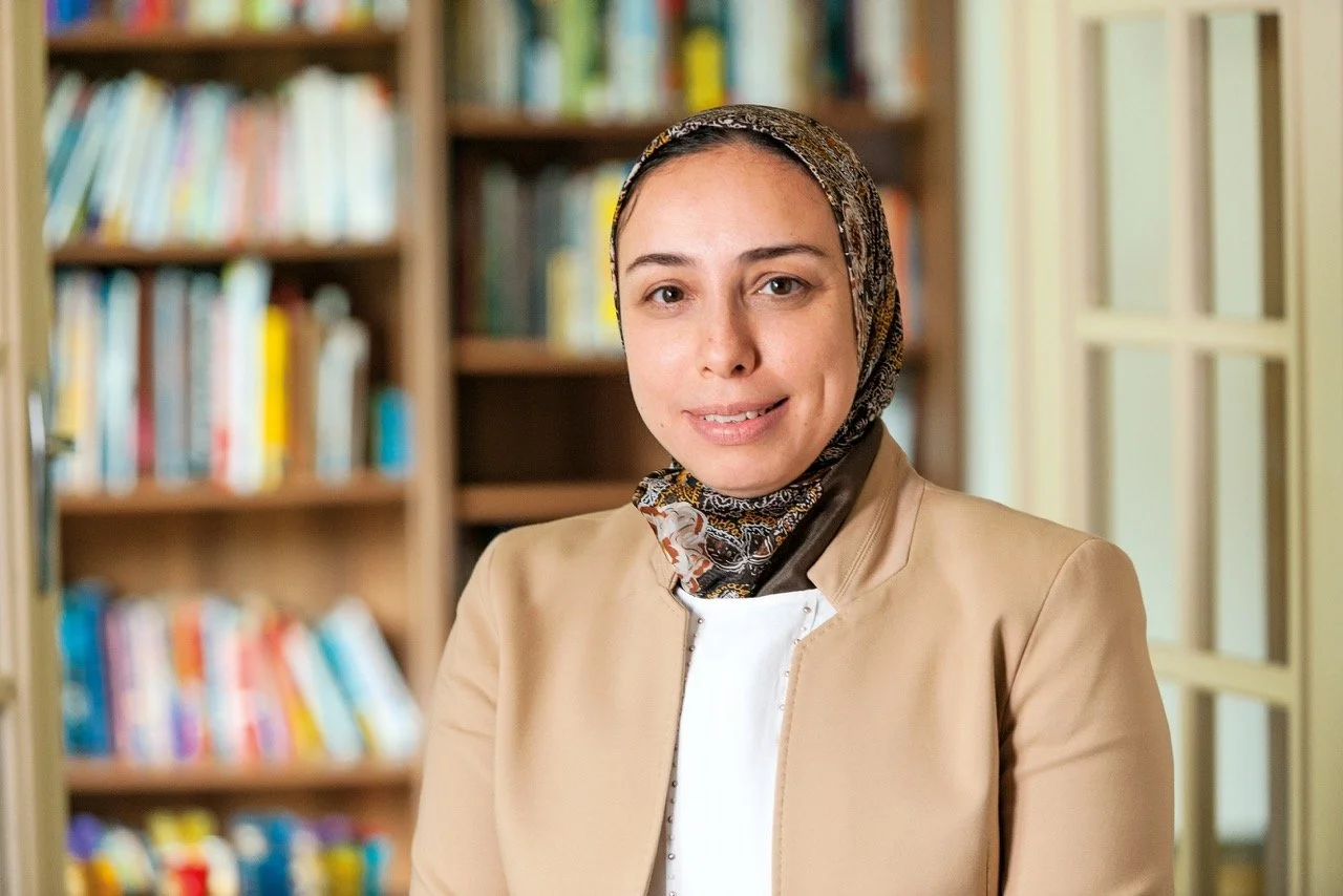 A woman wearing a patterned headscarf and beige blazer standing in front of a bookshelf filled with books.