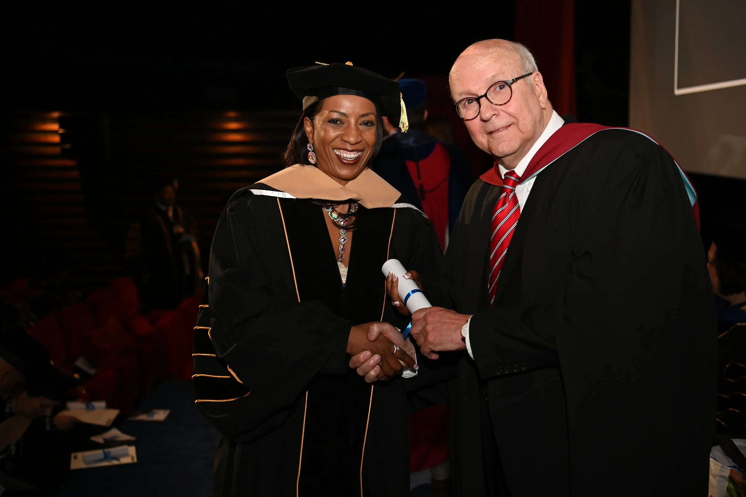 A woman and a man in academic regalia shaking hands at a graduation ceremony, with the woman holding a diploma.
