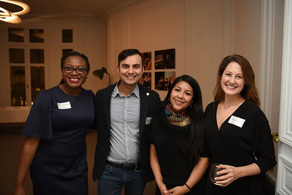 Four smiling people at a social event, standing close together indoors with framed pictures on the wall behind them.