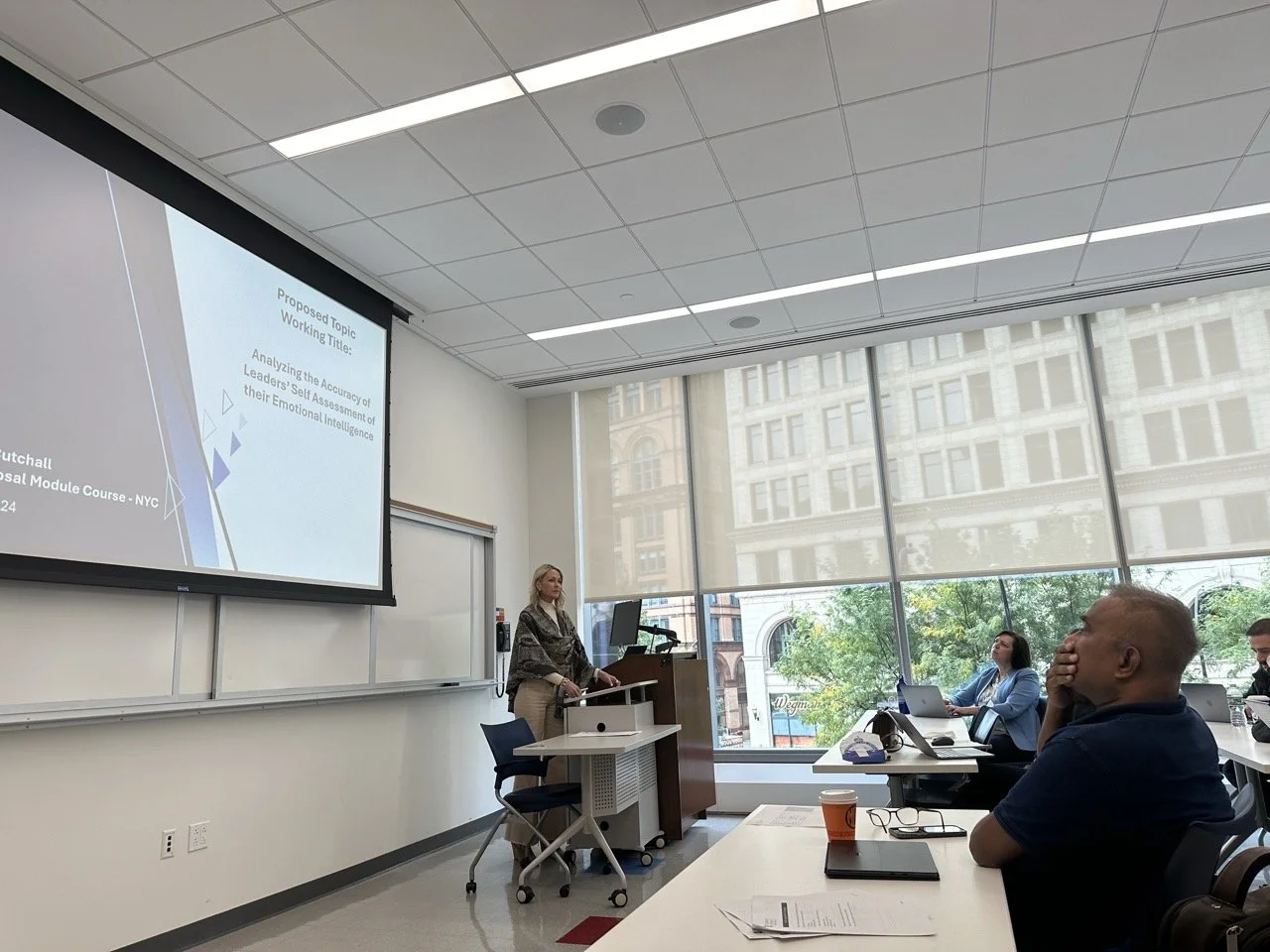 A woman giving a presentation in a classroom with large windows and a cityscape view. Several students are seated at desks, listening.