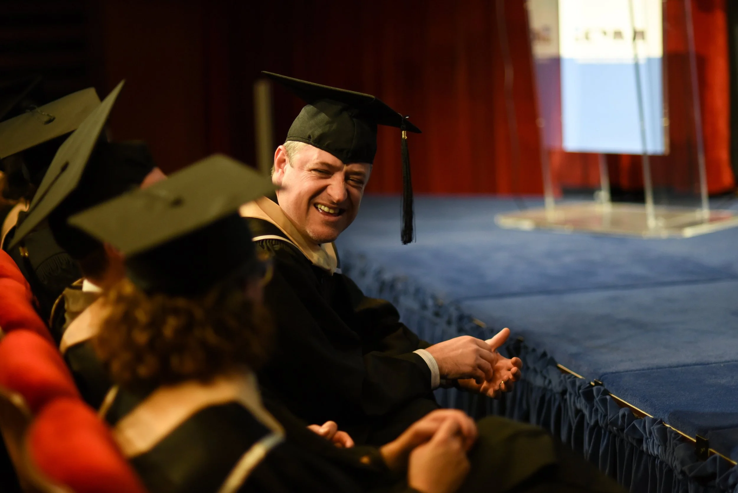 Graduates in black caps and gowns sitting in an indoor auditorium, smiling and laughing, with a stage in the background.