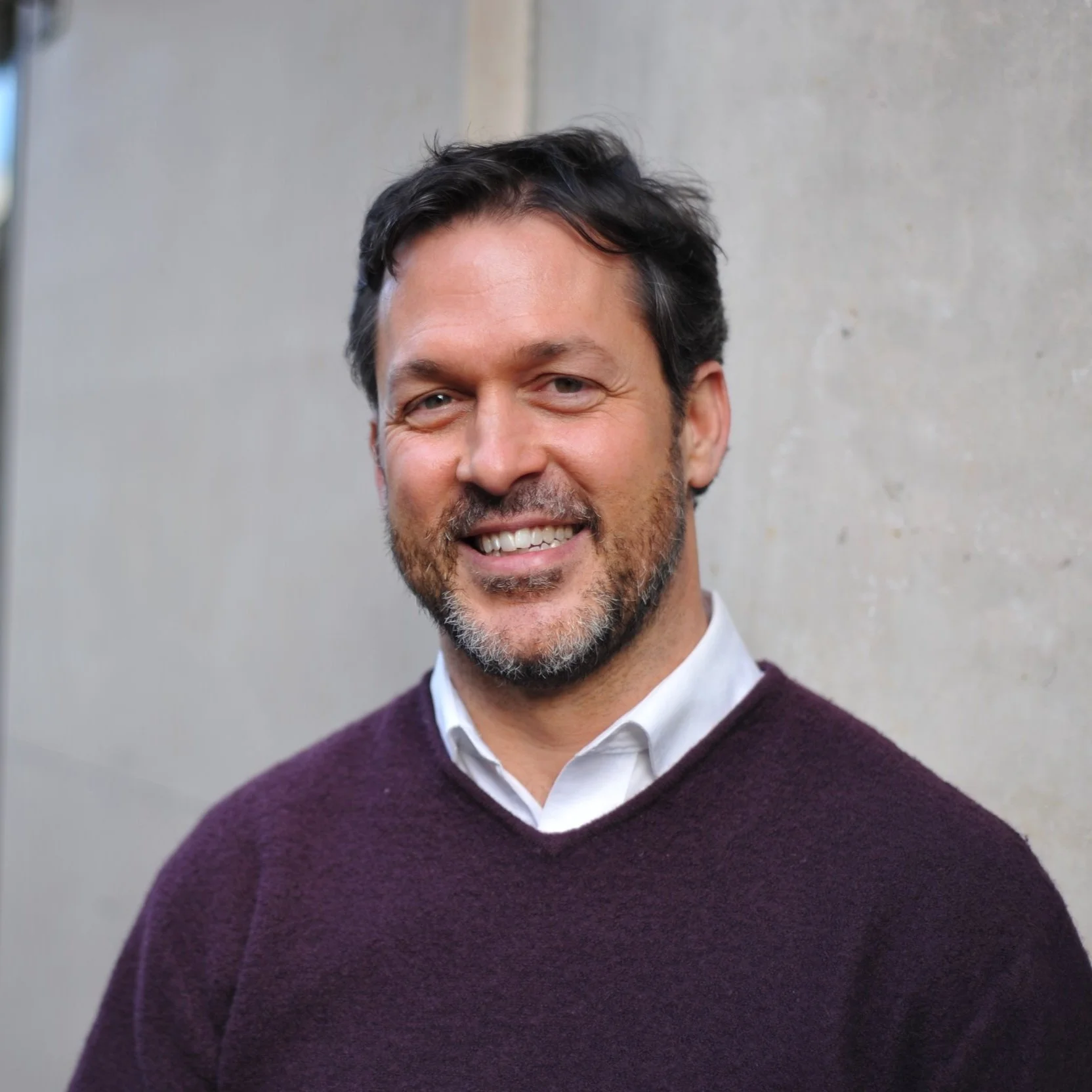 A smiling man with dark hair and a beard, wearing a white shirt under a purple sweater, standing against a neutral gray wall.