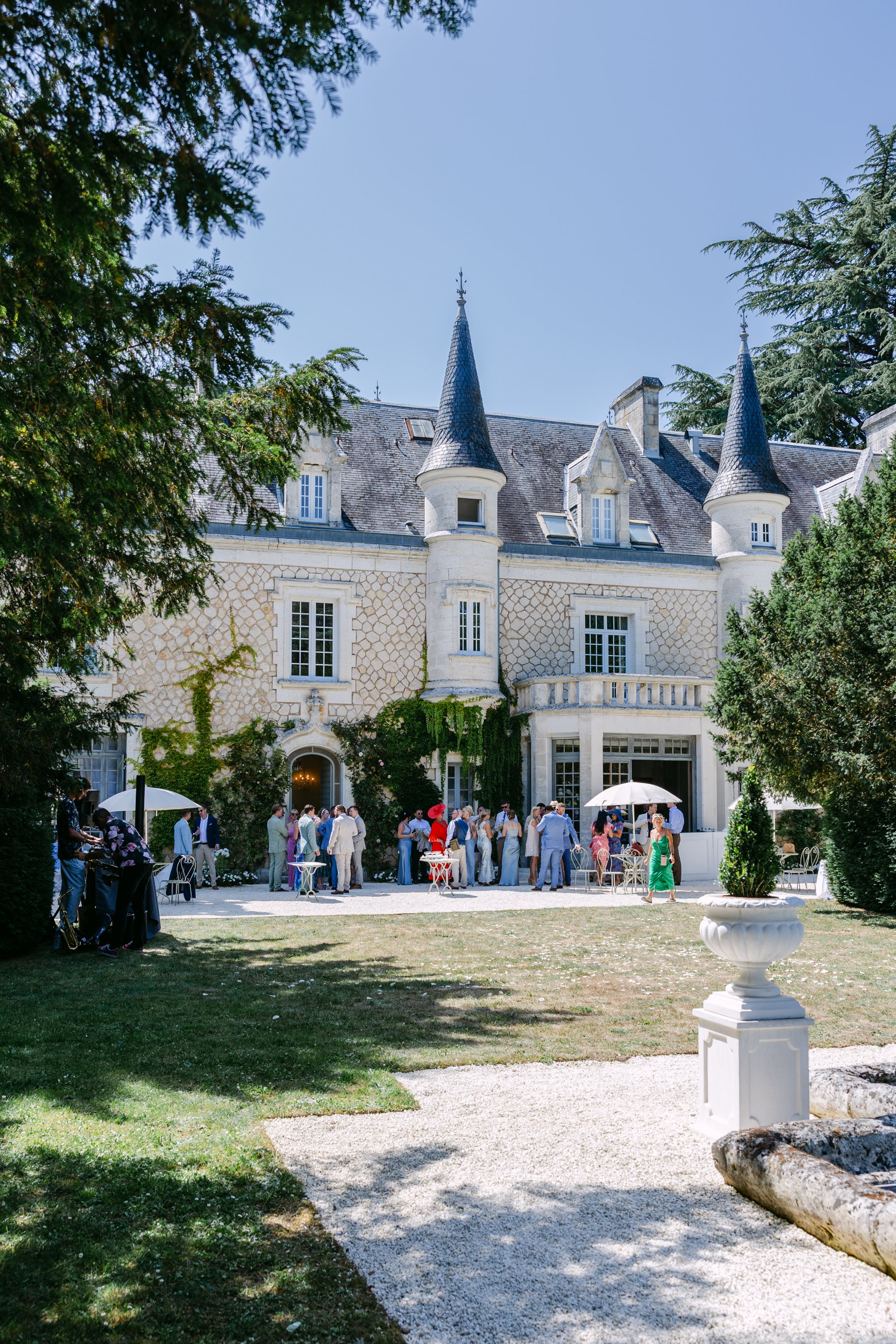 People gathered outside a castle-like building with towers, having an outdoor event on a sunny day.