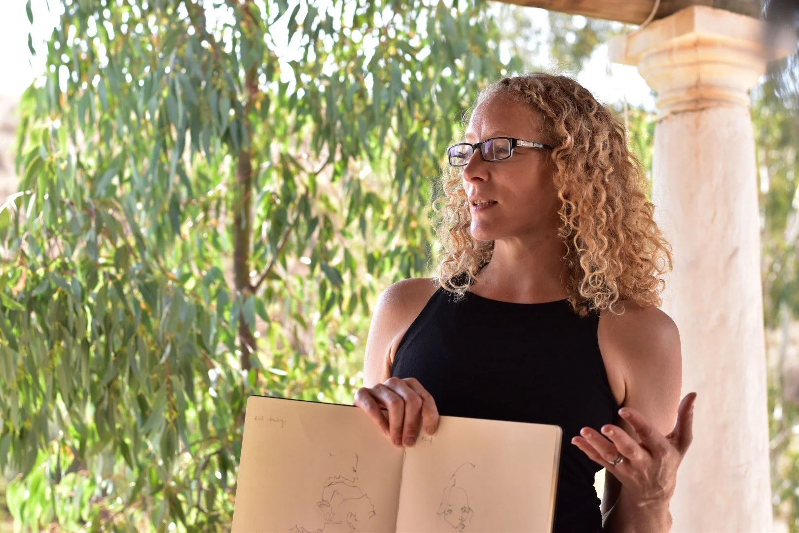A woman with curly blonde hair, wearing glasses and a sleeveless black top, is holding an open sketchbook and speaking outdoors with green foliage and a stone column in the background.