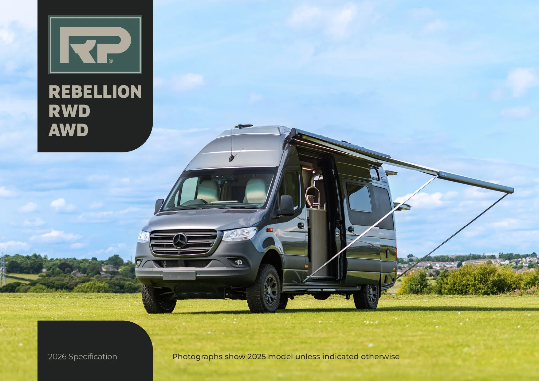 A silver camper van with an extended awning, parked on a grassy field with a blue sky and scattered clouds in the background.