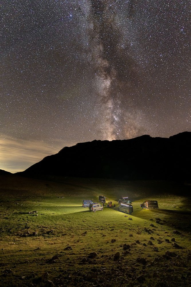 Nighttime scene of a starry sky with the Milky Way visible above dark mountain silhouettes and a grassy field where several vehicles are parked, and a few people are gathered around illuminated spots.