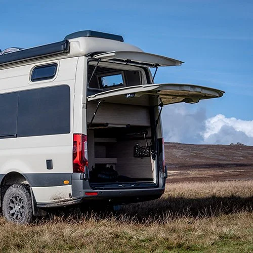 Open van with ceiling hatch and side panels, equipped for work or storage, parked on grass with cloudy sky in the background.