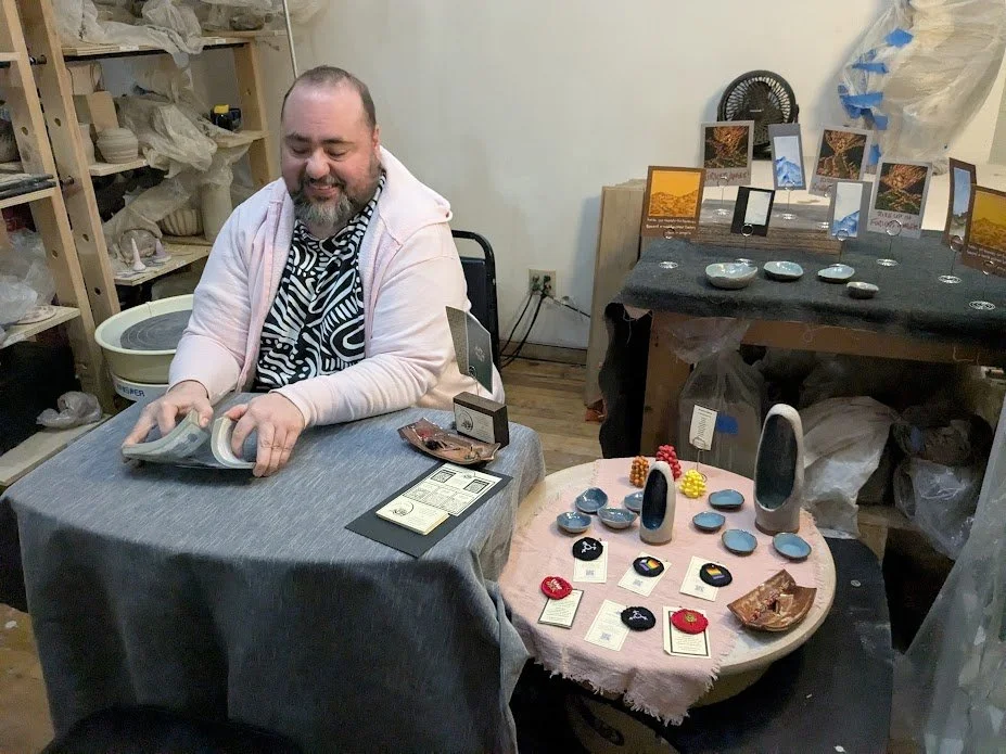 owen is wearing a pink hoodie over a black and white t-shirt, shuffling cards at a table covered with a blue-gray cloth. To one side is a smaller round table covered with a pink cloth, displaying Owen's art