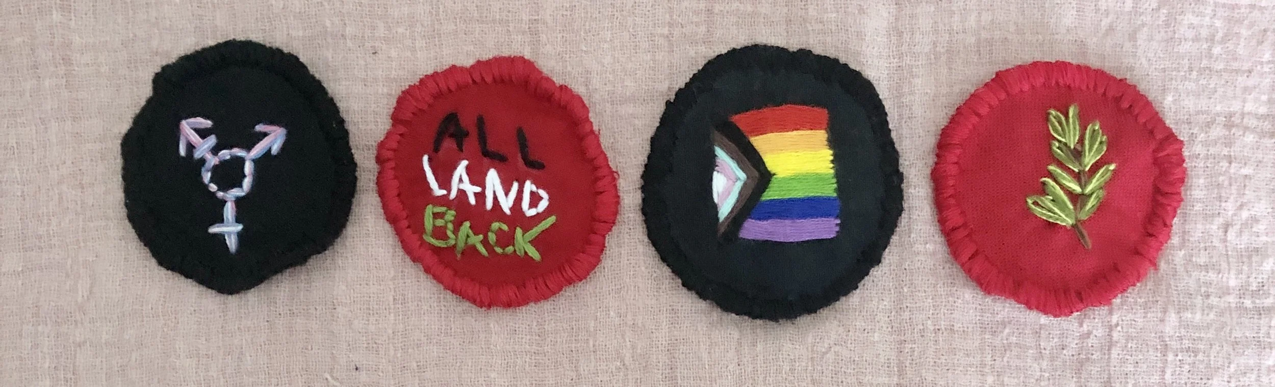 4 prayer pouches in a row, a trans pride symbol on black, "all land back" on red, rainbow progress flag on black, and a rosemary sprig on red fabric. The pouches are embroidered in the shape of circles