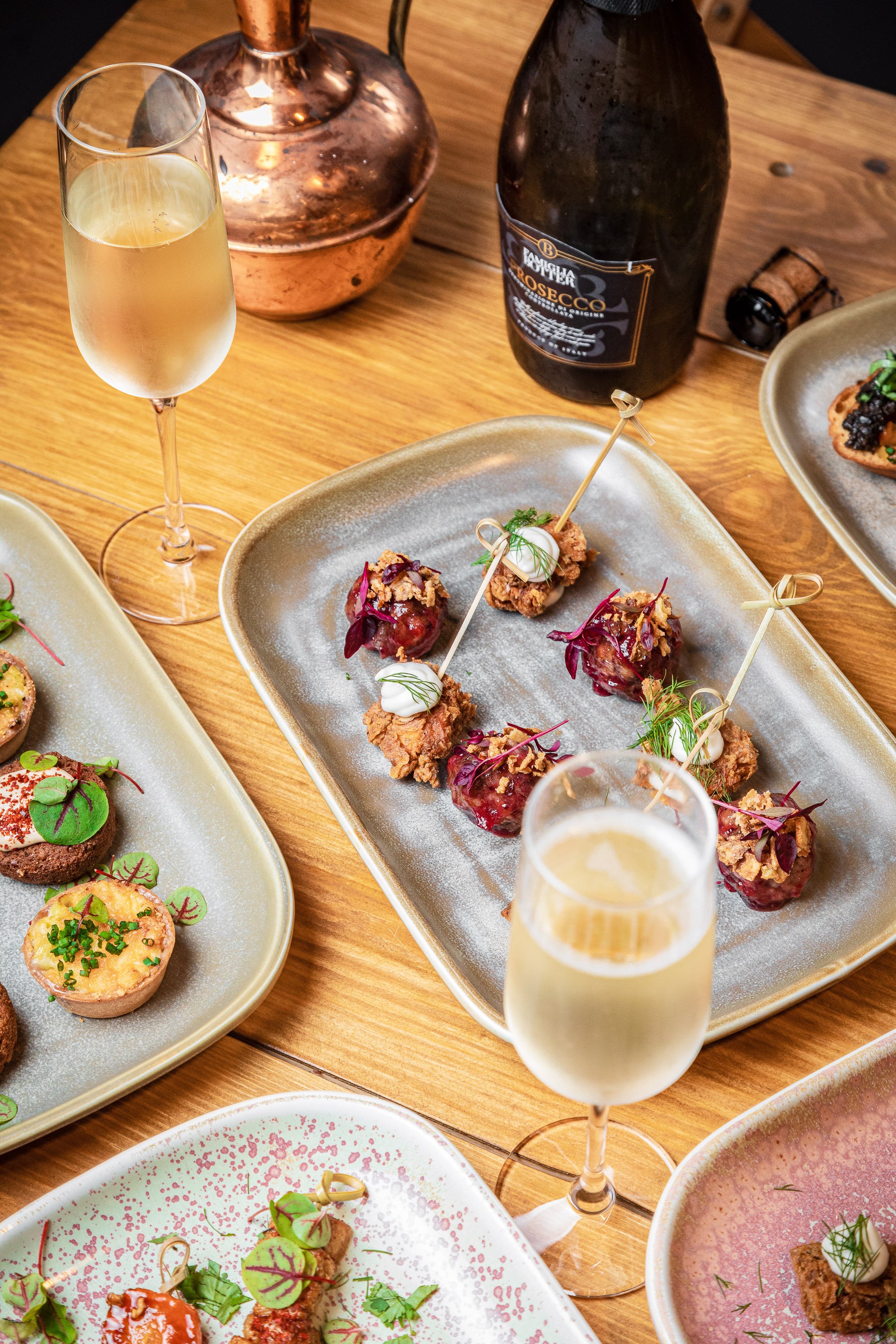 A table with plates of assorted appetizers accompanied by two glasses of champagne and a bottle of sparkling wine served at 'The Stables'