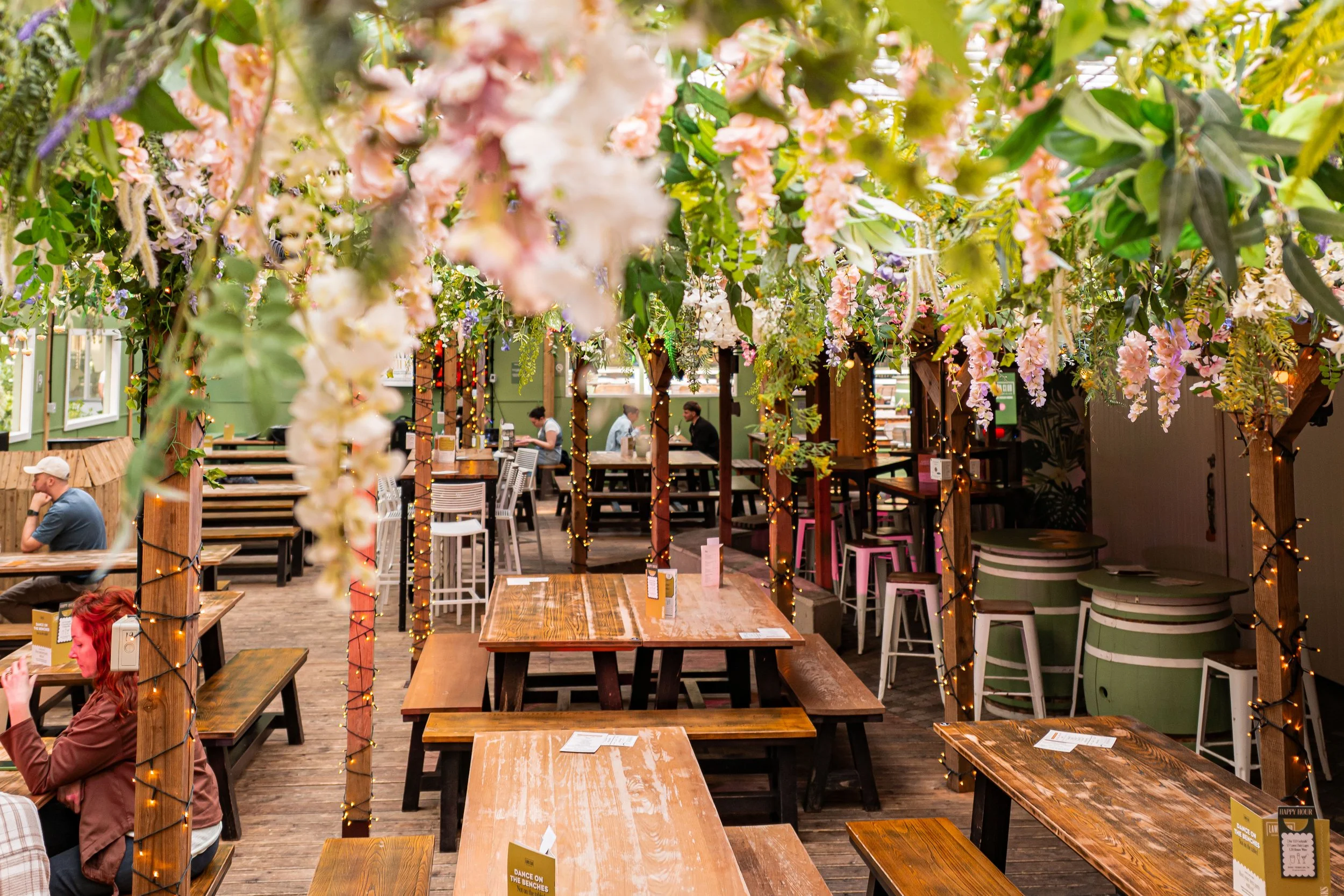Bar seating area filled with wooden picnic tables. The ceiling is adorned with hanging plants and foliage creating a stylish and cosy vibe.