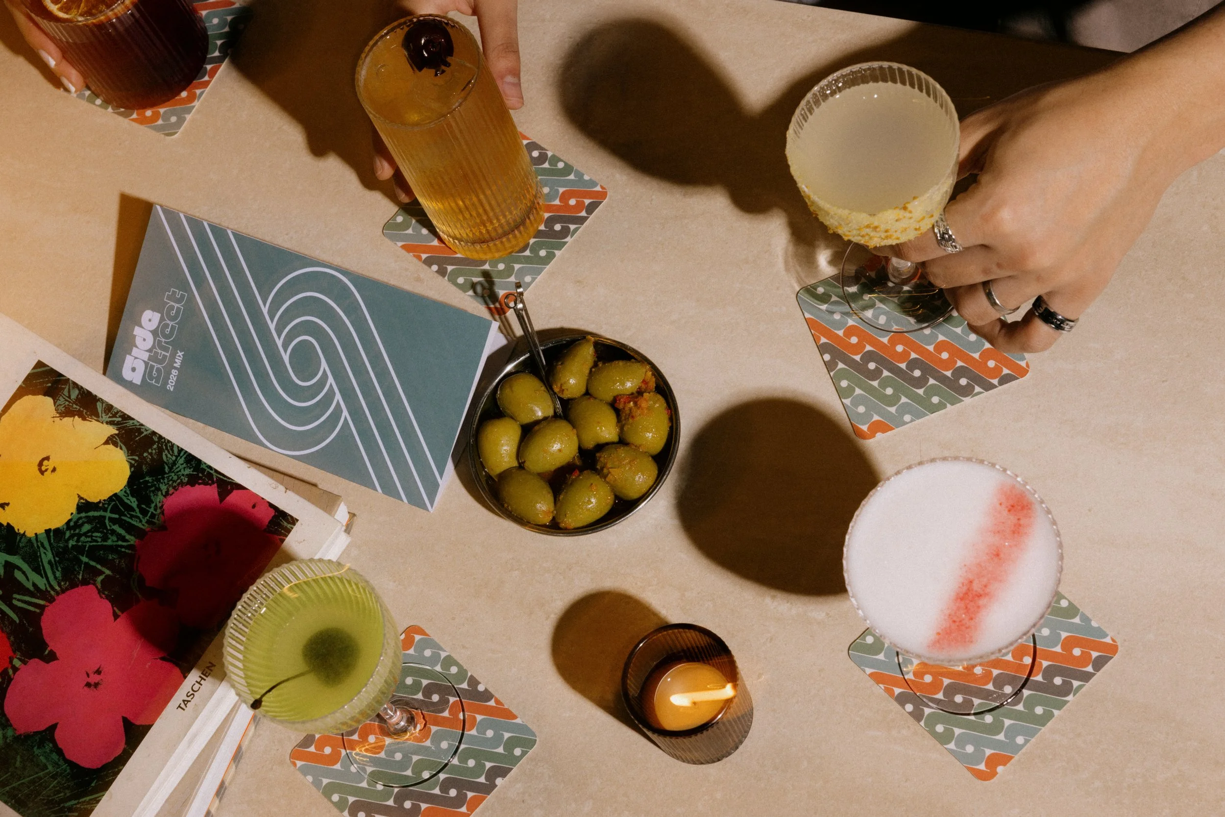 Top view of a table with several drinks, including champagne in flutes and a cocktail with a cherry on top, along with bowls of green olives and some party decorations and papers.