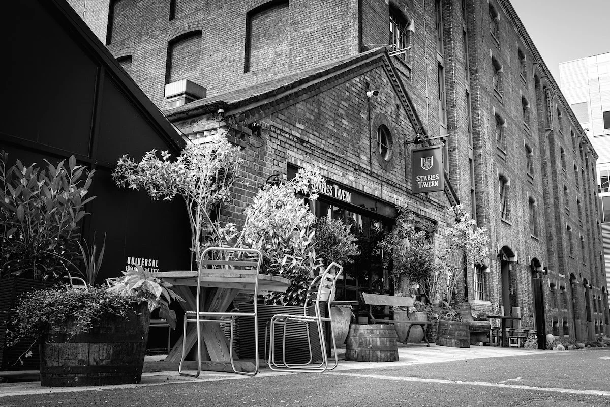 Black and white photo of outside of a brick building with potted plants, tables, and chairs in front of a tavern named 'Stables Tavern'.