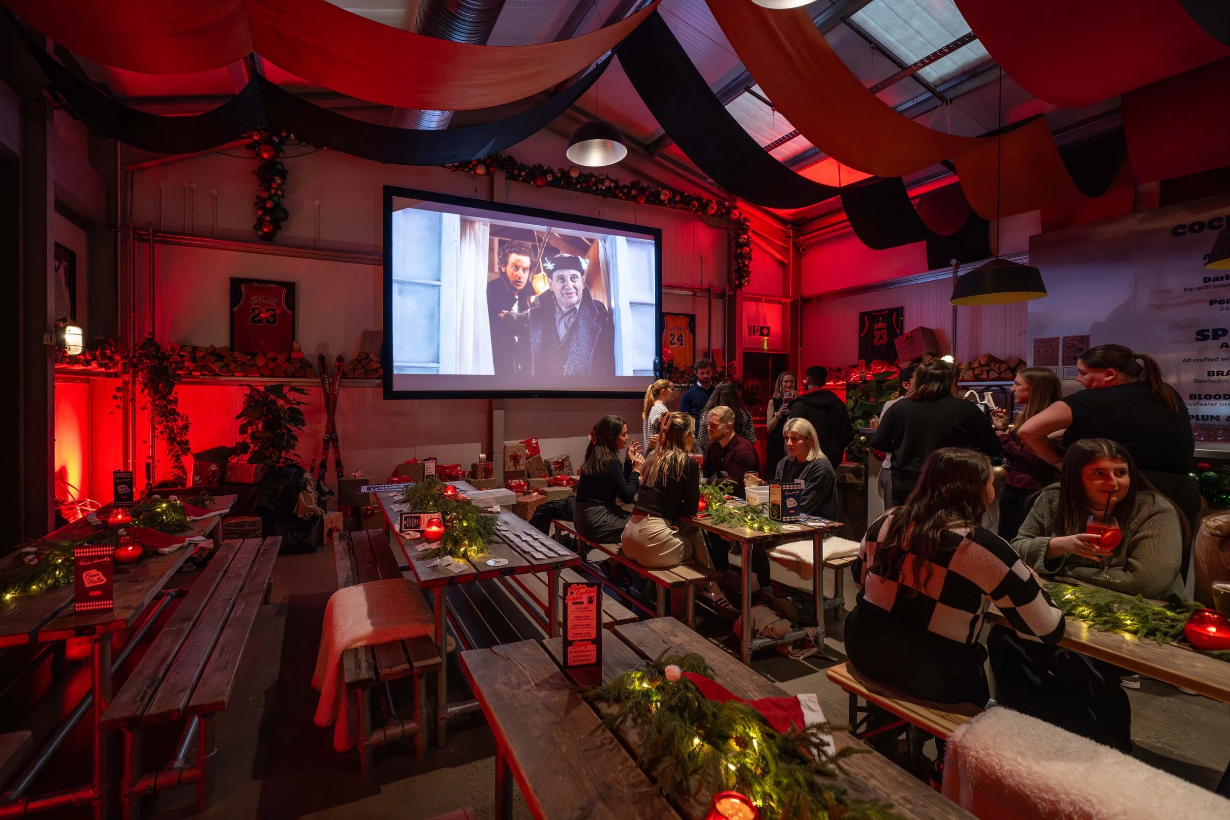 Interior of a modern bar or cafe with wooden tables and benches, a counter with staff and customers, a dark green accent wall decorated with framed pictures, and television screens showing sports