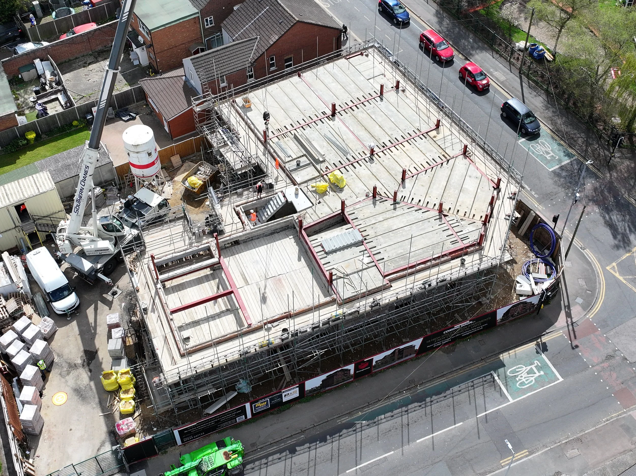 Aerial view of a construction site with scaffolding, concrete flooring, and construction vehicles, located in an urban area with nearby parked cars, buildings, and a bike lane on the street.
