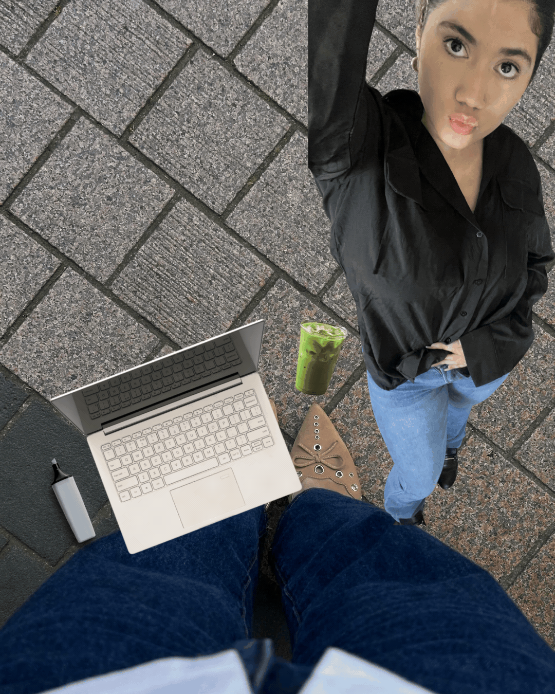 From a first-person perspective, a person is standing on a brick sidewalk holding a small laptop with a virtual keyboard, with a pen and a green iced drink nearby. In front, a woman with dark hair tied back, wearing a black jacket and blue jeans, looks up at the camera with puckered lips.