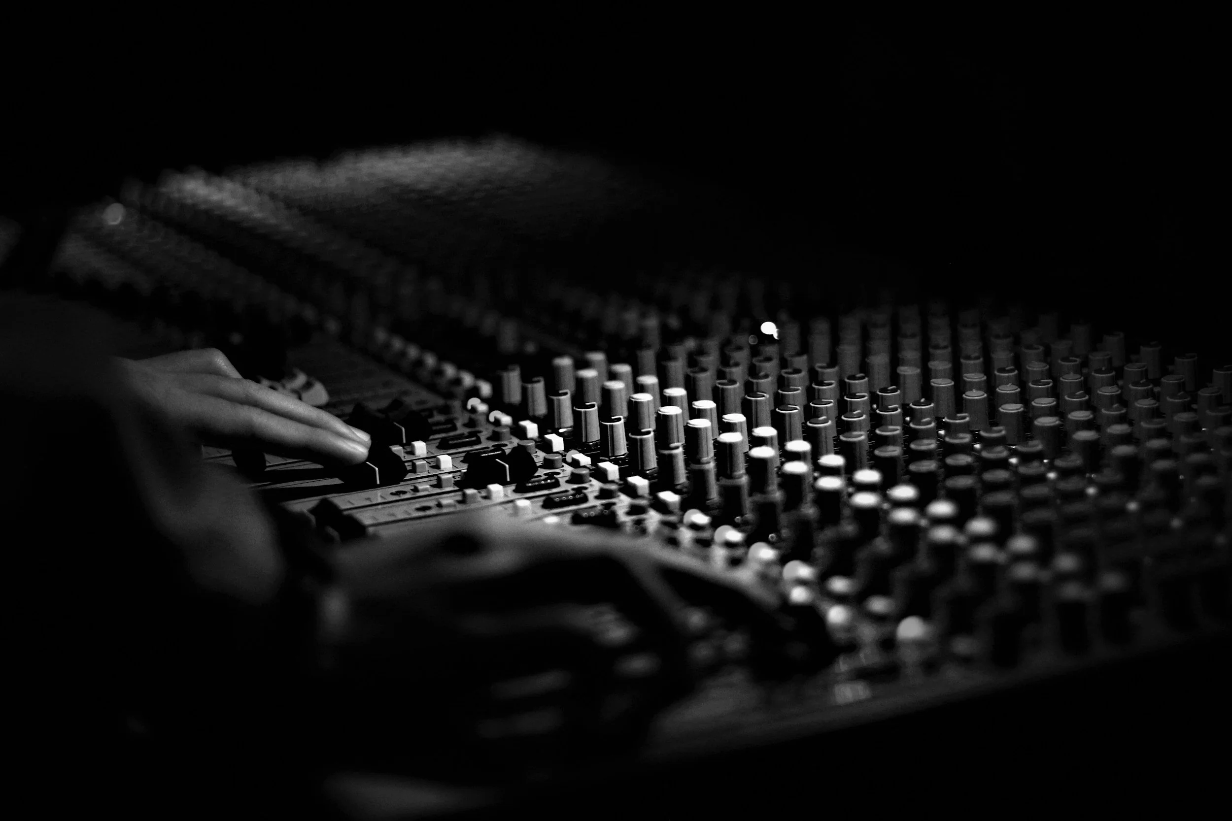 Black and white photo of hands controlling a sound board's knobs and sliders