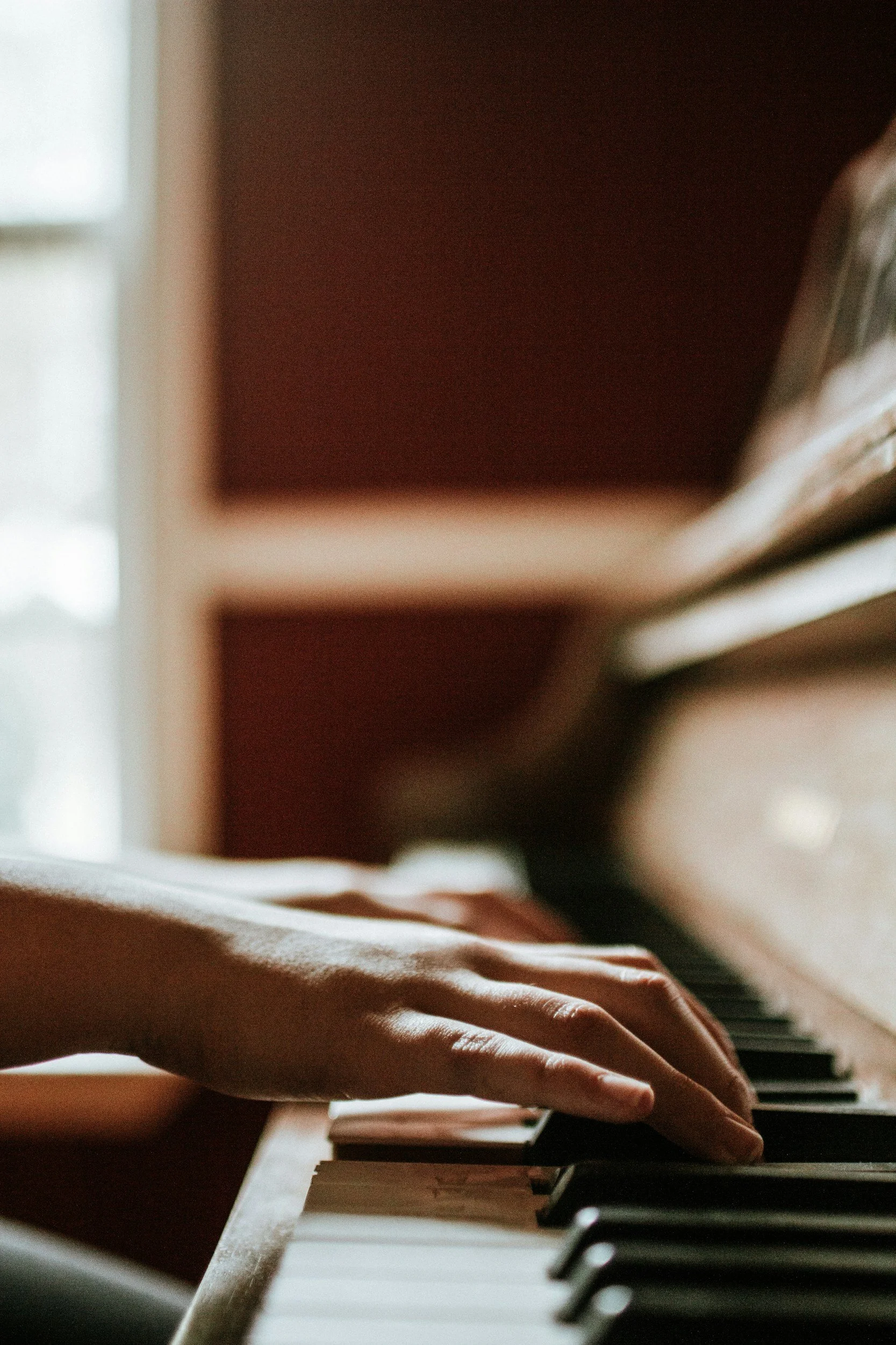 Close-up of a person's hand playing the black and white keys on a wooden piano, with a window in the background.