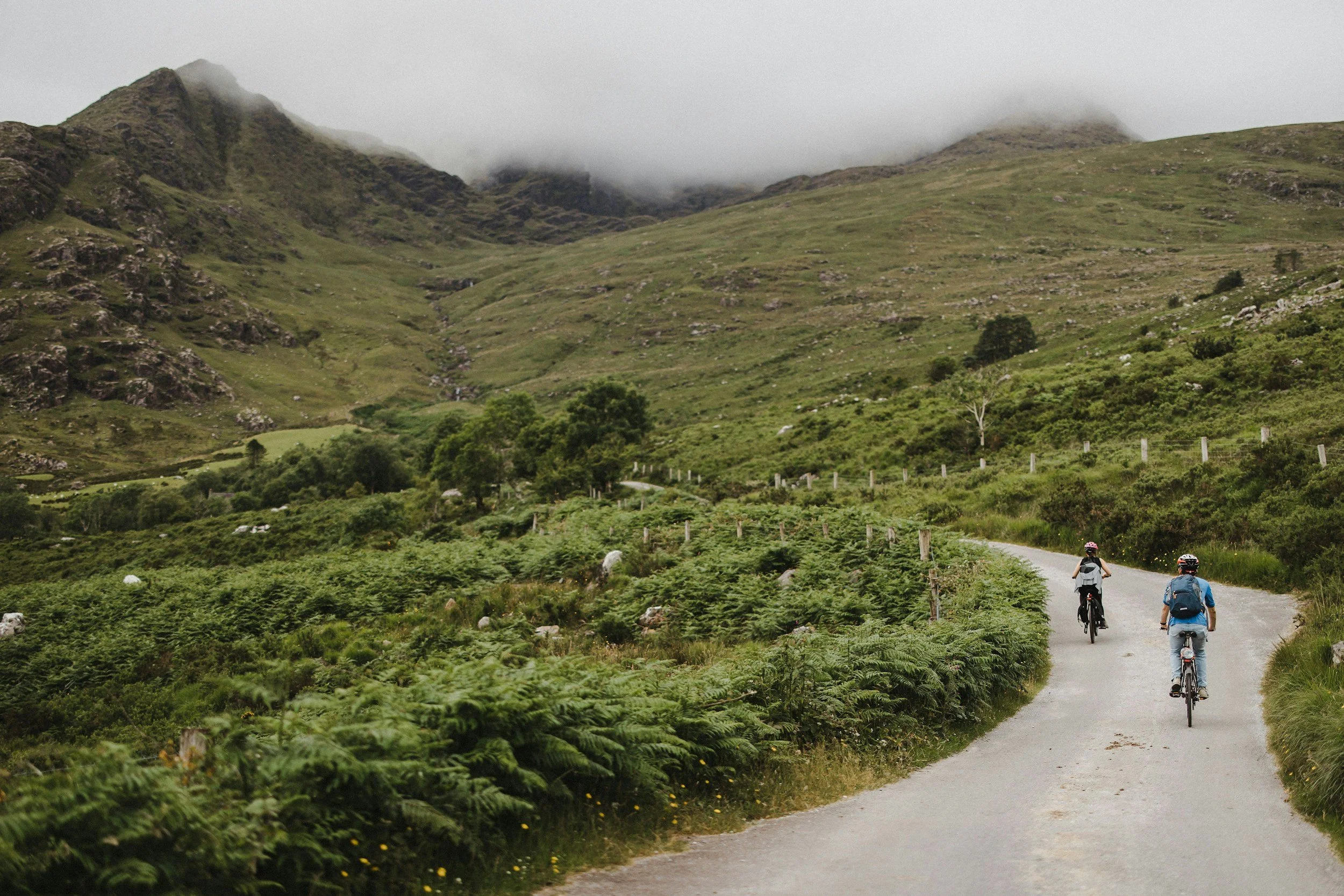 Two cyclists riding on a narrow road through a lush green valley with mist-covered mountains in the background.