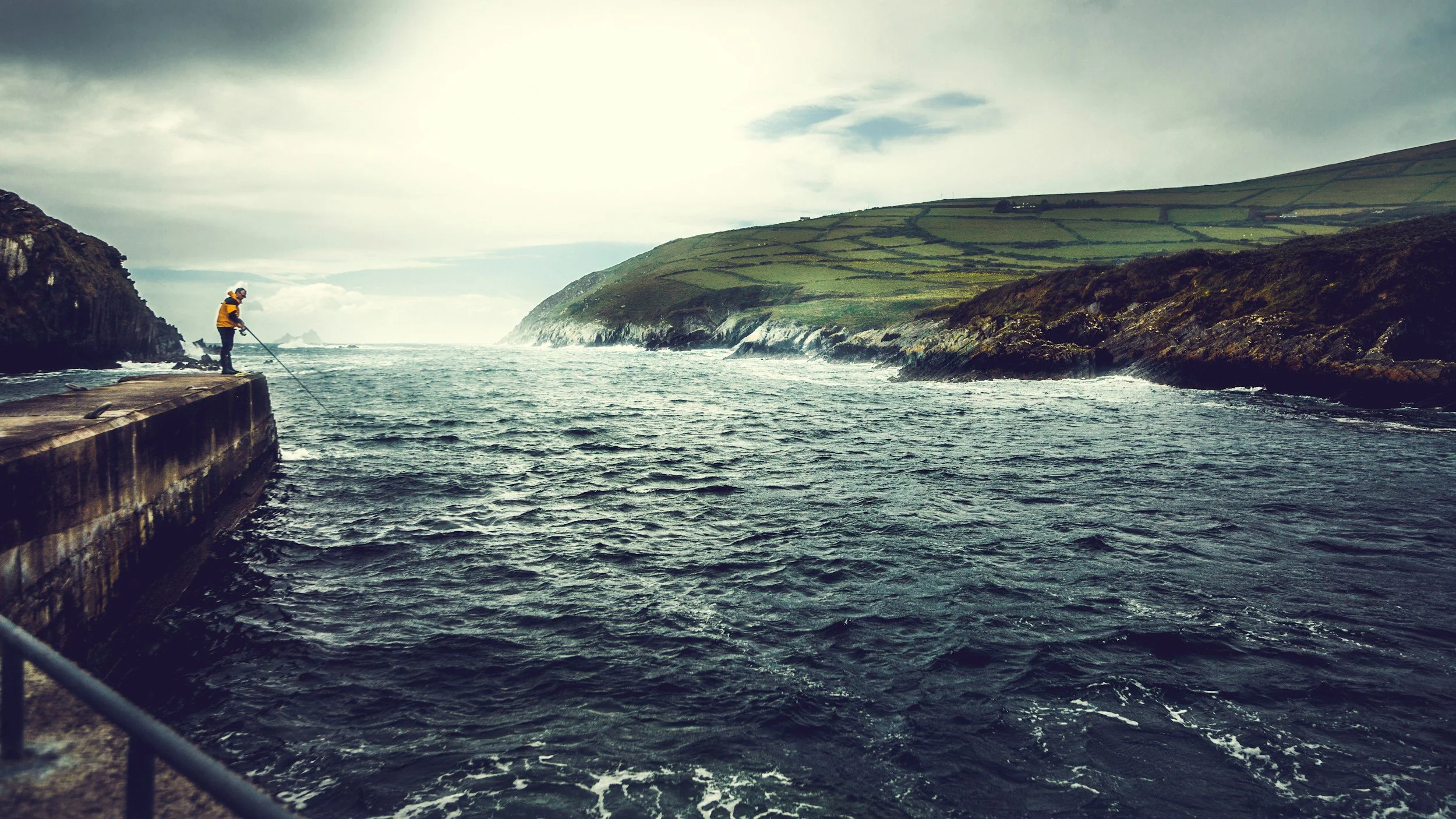 Person fishing on a pier with rocky coastline and ocean waves in the background.