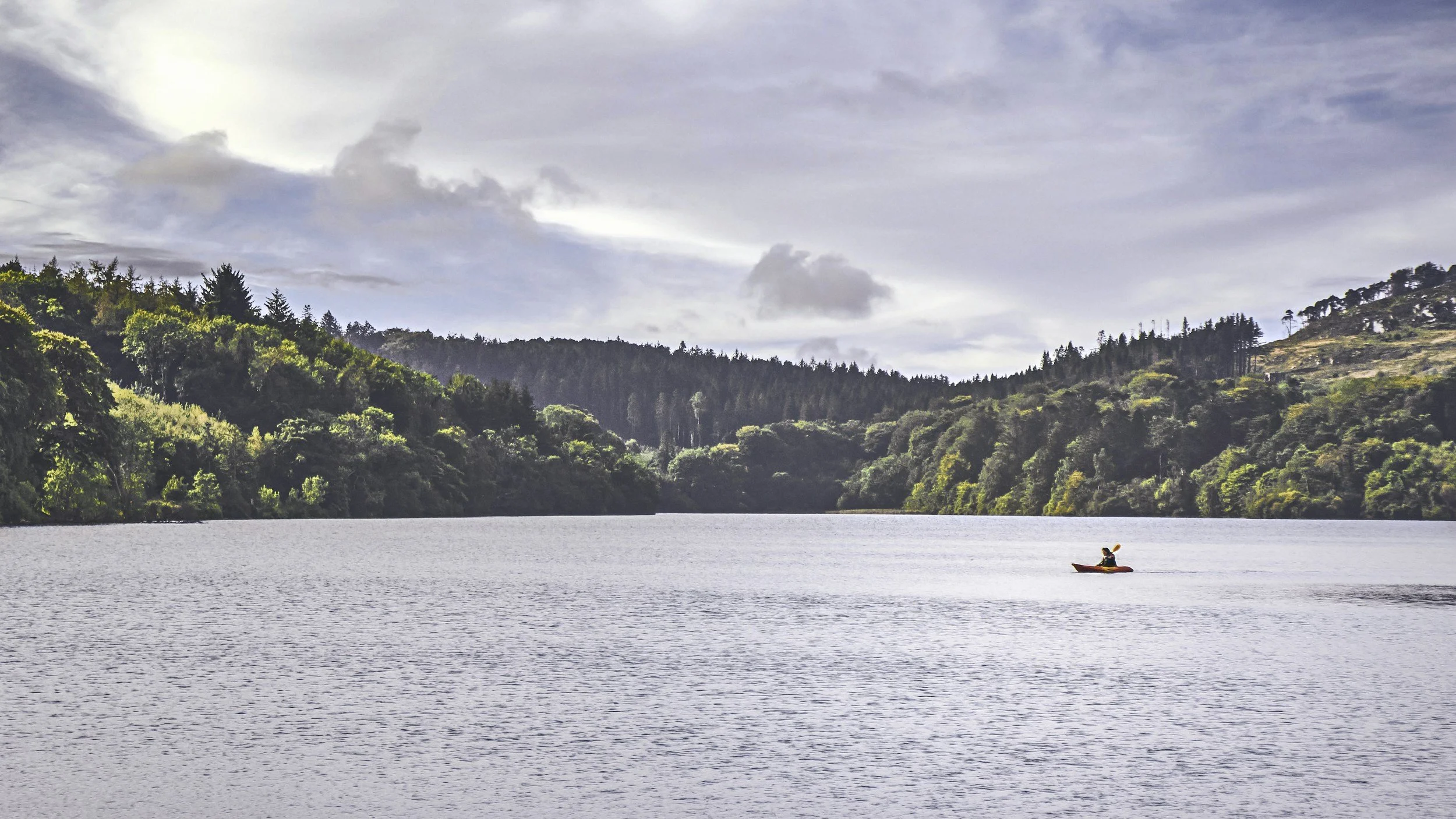A person kayaking on a lake surrounded by forested hills under a cloudy sky.
