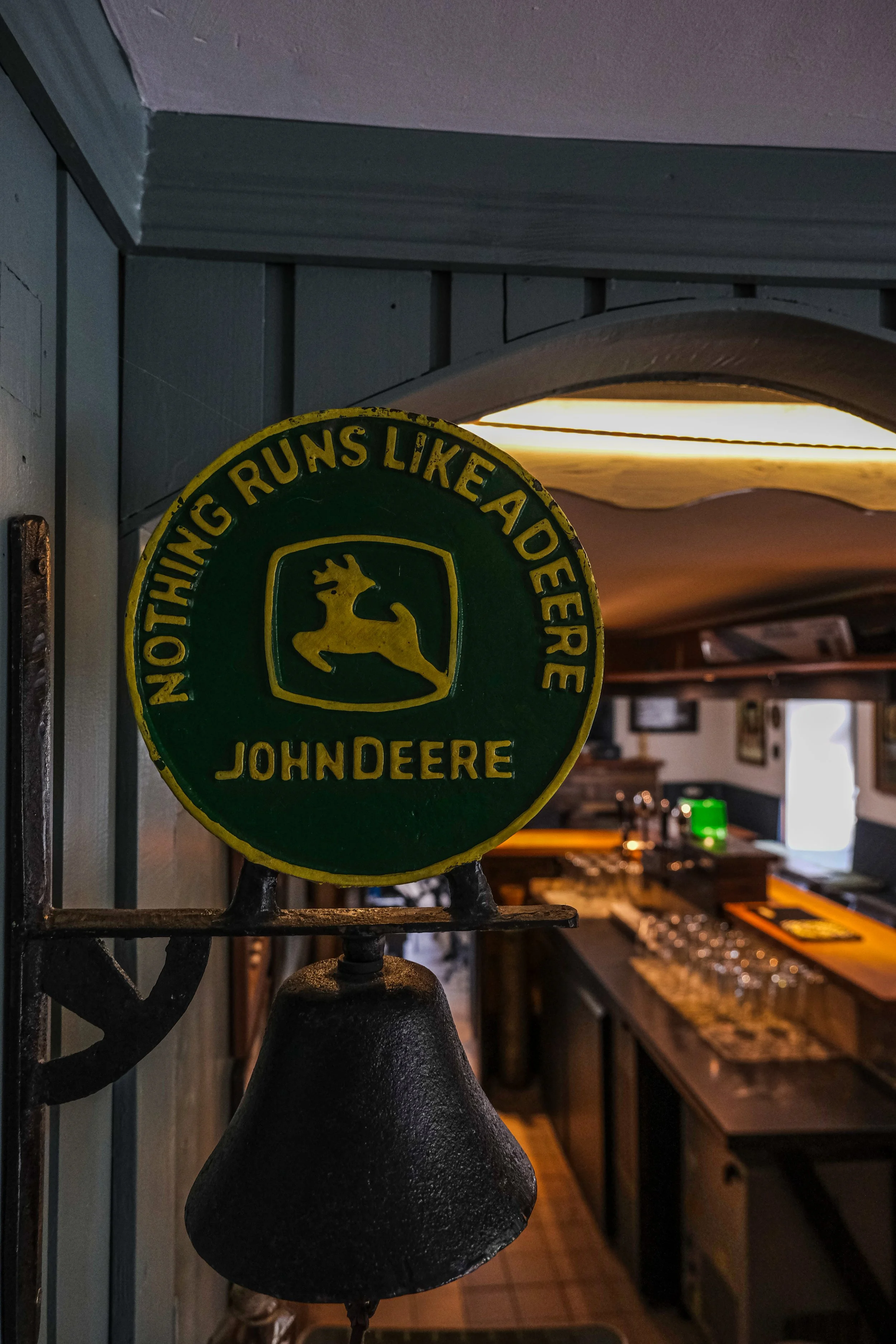 A vintage John Deere sign with the slogan "Nothing runs like a Deere" and a bell, inside a dimly lit bar.