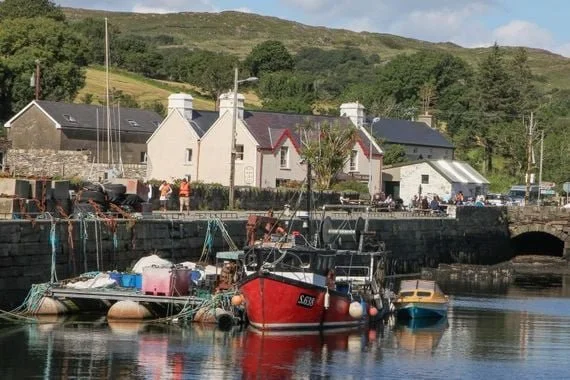 Scenic harbor with boats docked and quaint houses in the background, surrounded by lush greenery and hills.