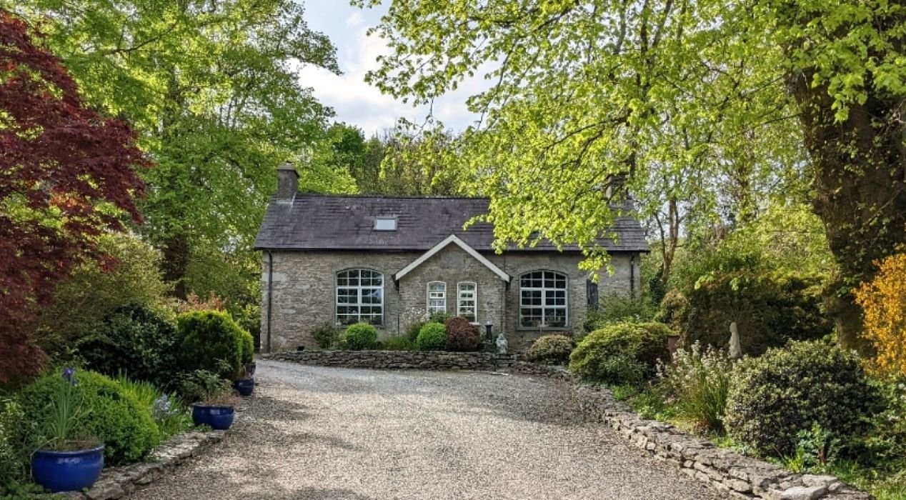 Charming stone cottage surrounded by lush greenery and gardens, with a gravel driveway leading to the entrance.