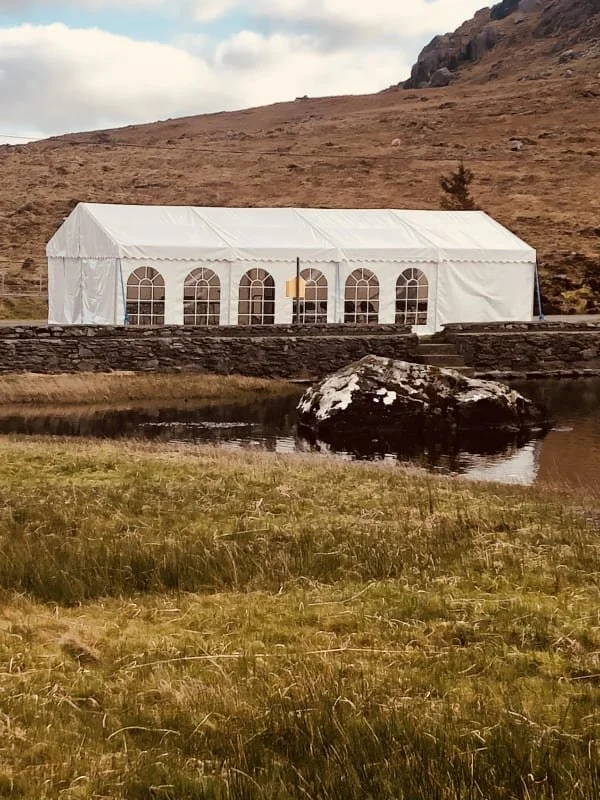 Large white event tent with arched windows situated by a small pond on grassy terrain with a rocky hillside and mountain in the background.