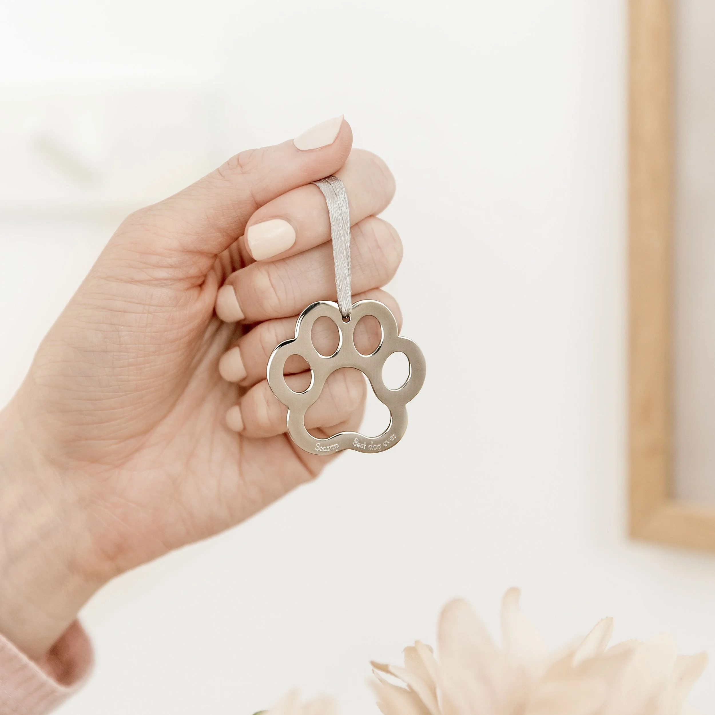 A hand holding a silver pet tag shaped like a paw print, attached to a gray ribbon.