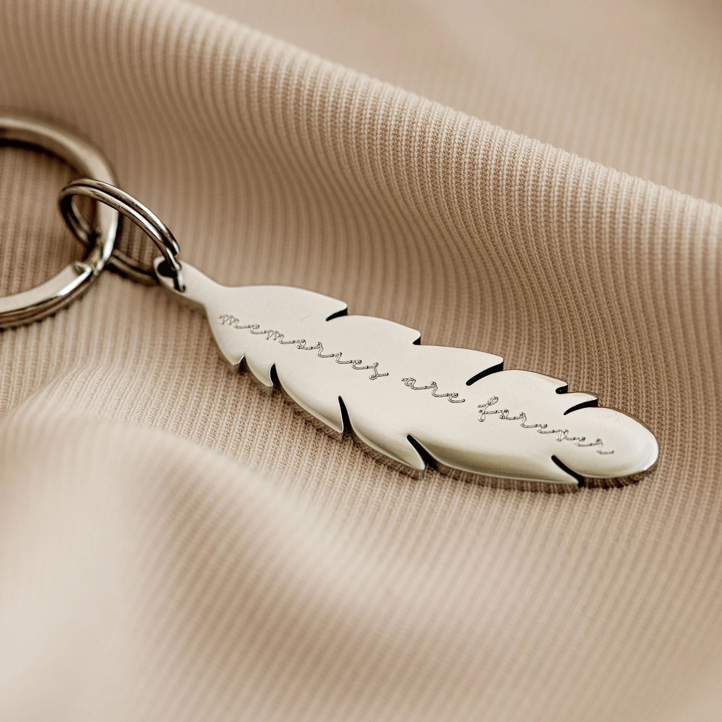 Close-up of a silver feather-shaped keychain with an engraved message, resting on soft, beige fabric.